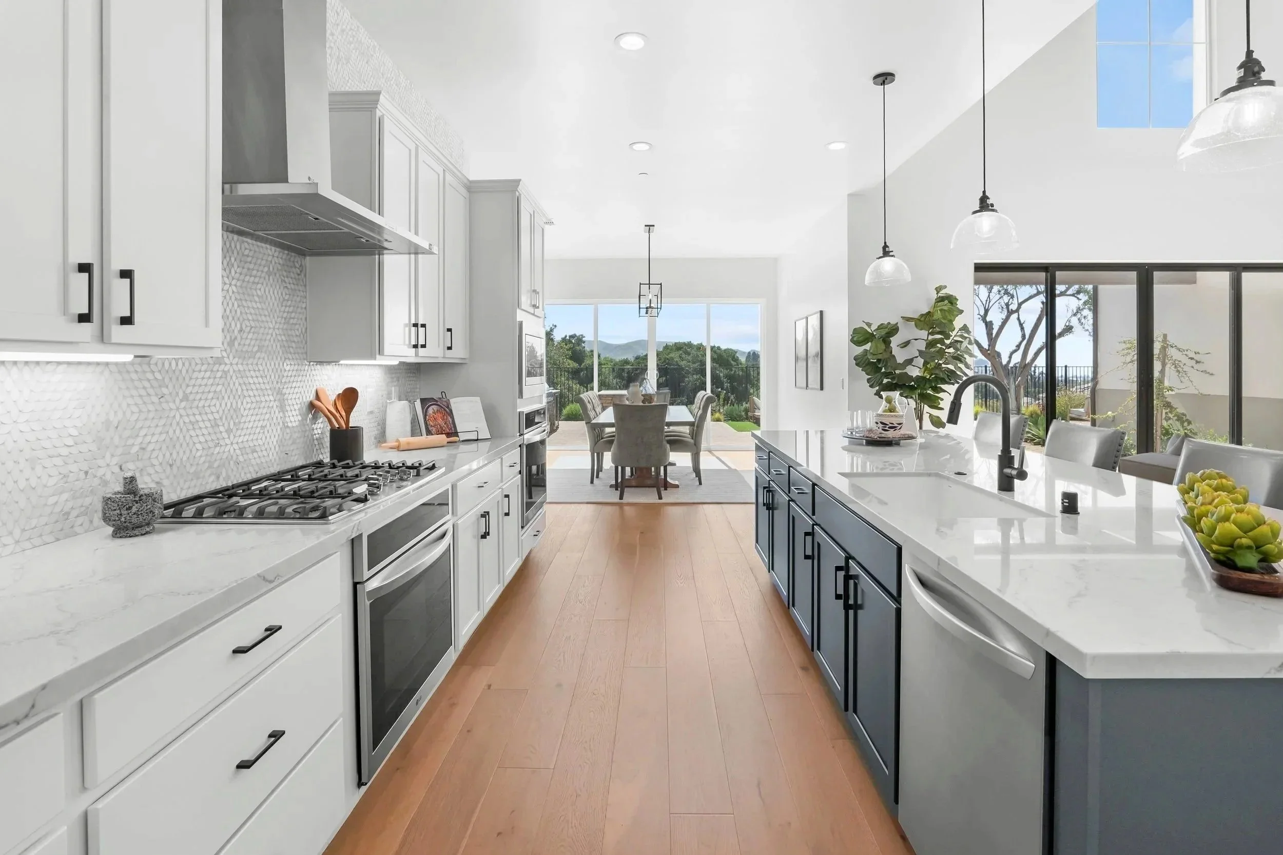 Modern kitchen with white and gray cabinets, marble countertops, wood flooring, and a large island with bar seating. There are three pendant lights hanging above the island, a leafy plant, and a view of a dining area and outdoor landscape through large windows.