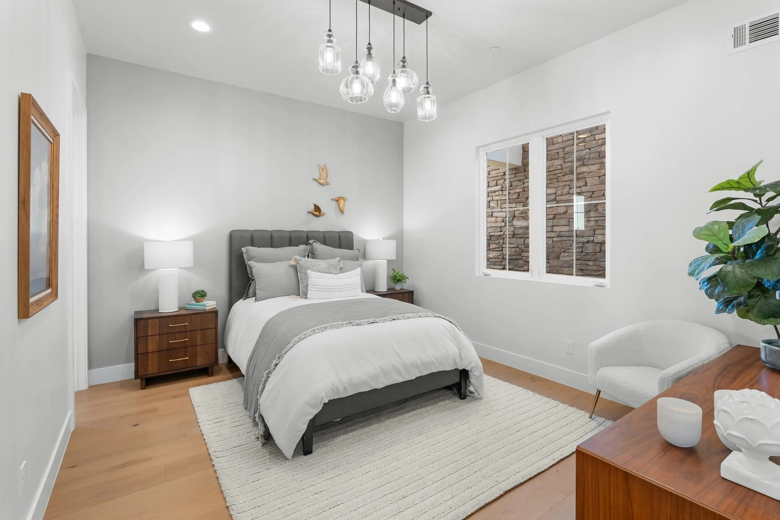 A modern bedroom with a gray upholstered bed, white and gray bedding, wooden nightstands with white lamps, a textured white rug, a window with a brick wall outside, a potted plant, and a wooden dresser with decor items.