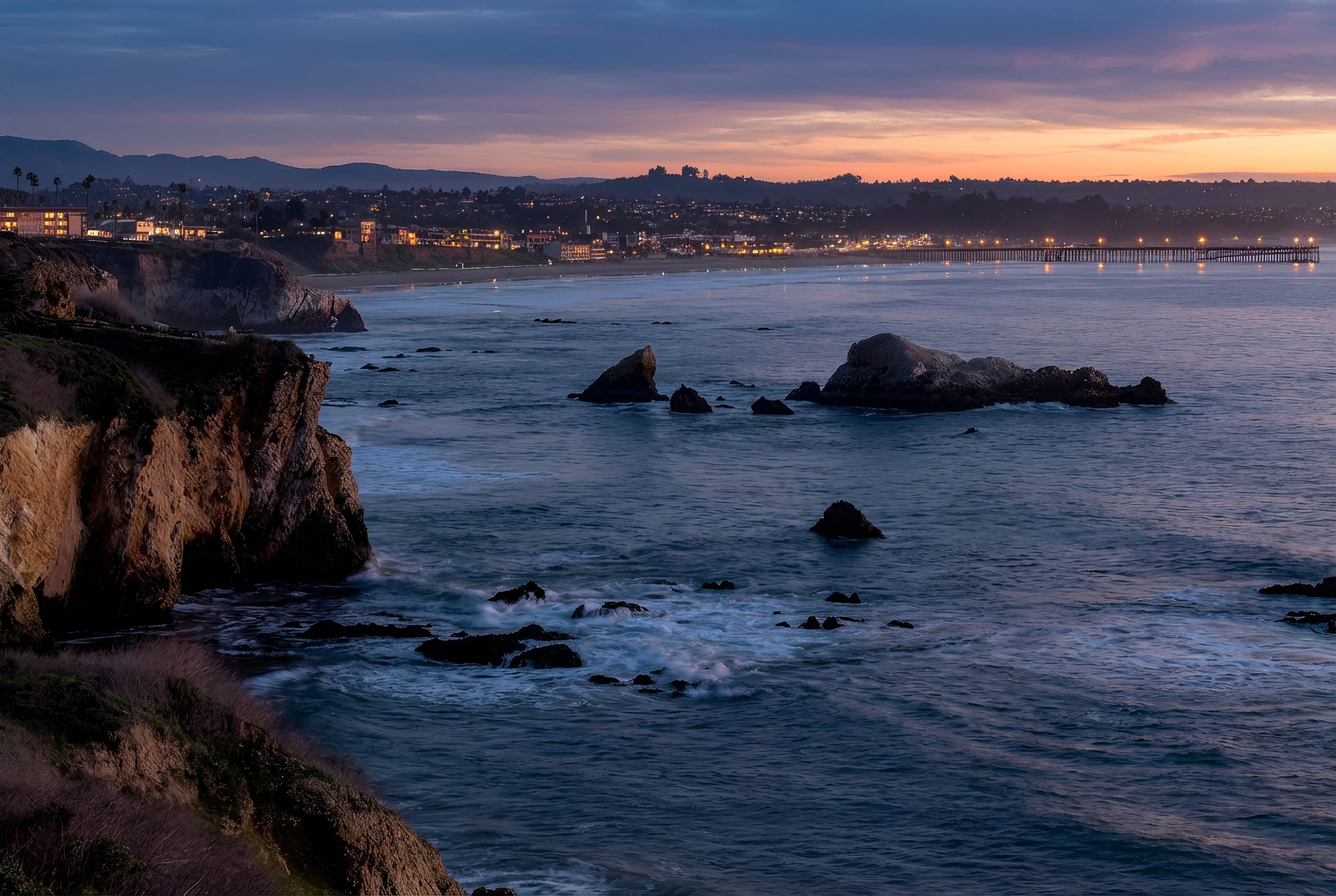 Sunset over coastal cliffs and ocean in San Diego, California with city lights and a pier in the distance.