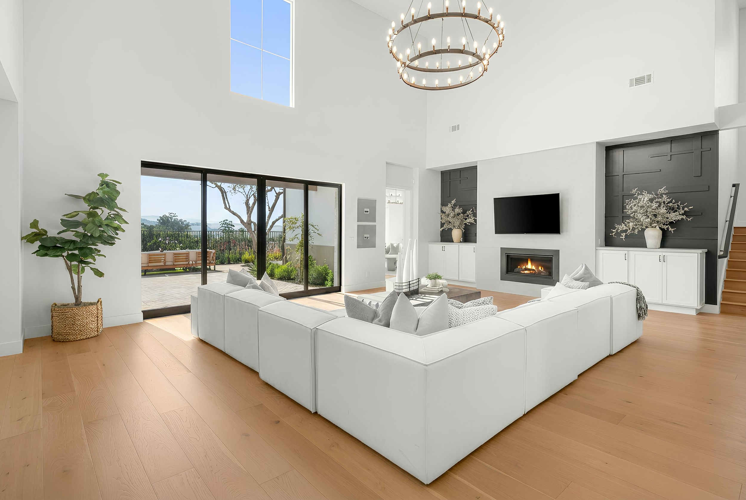 Bright living room with white sectional sofa, large sliding glass doors opening to outdoor patio with seating, fireplace, flat-screen TV, chandelier, and decorative vases with flowers.