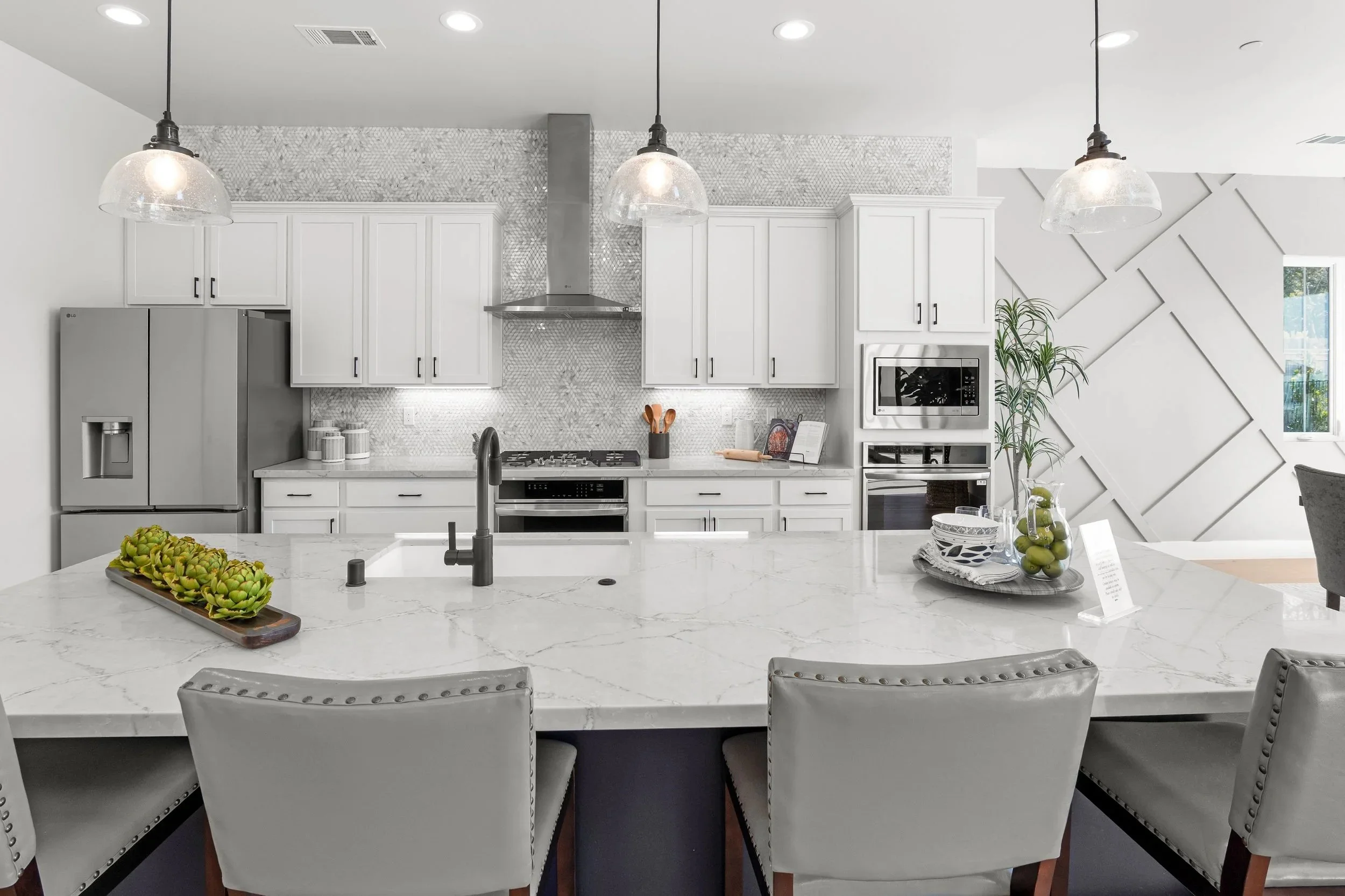 Modern white kitchen with marble island, stainless steel refrigerator, oven, microwave, and decorative wall paneling, with plants and tableware.