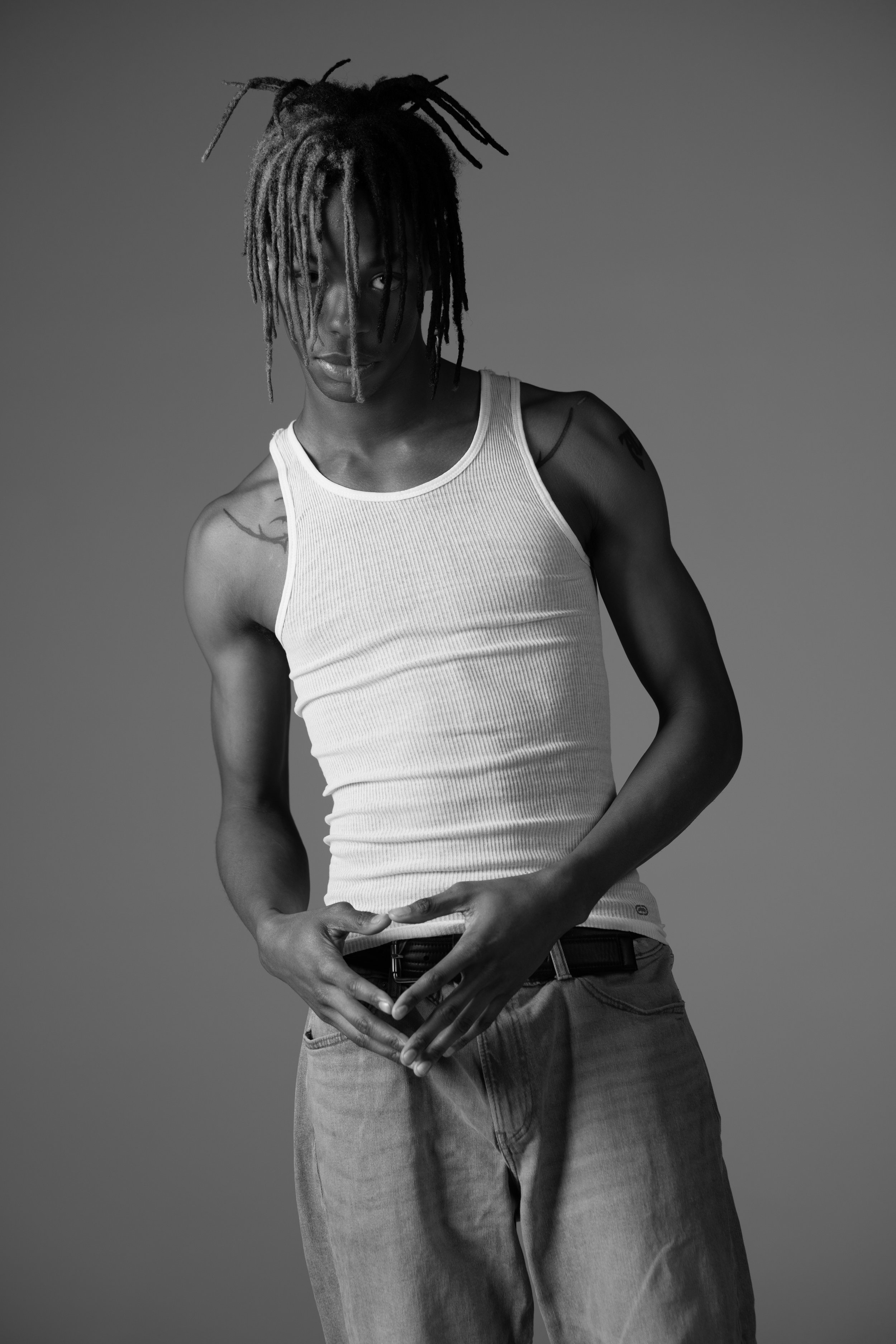 A young man with dreadlocks, wearing a sleeveless white tank top and jeans, stands against a plain background.