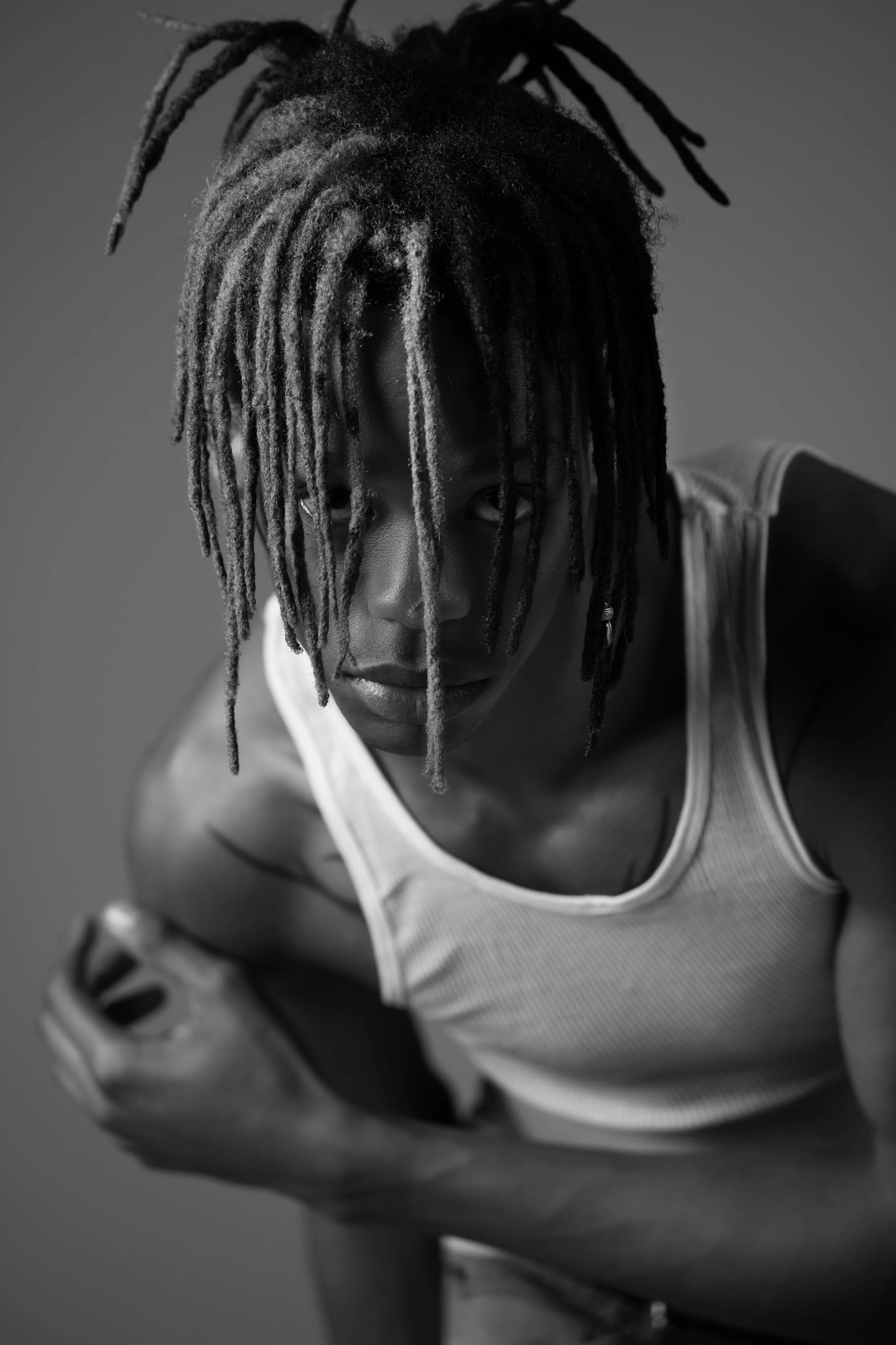 A black and white photo of a person with dreadlocks falling over their face, wearing a sleeveless shirt, looking towards the camera with a serious expression.