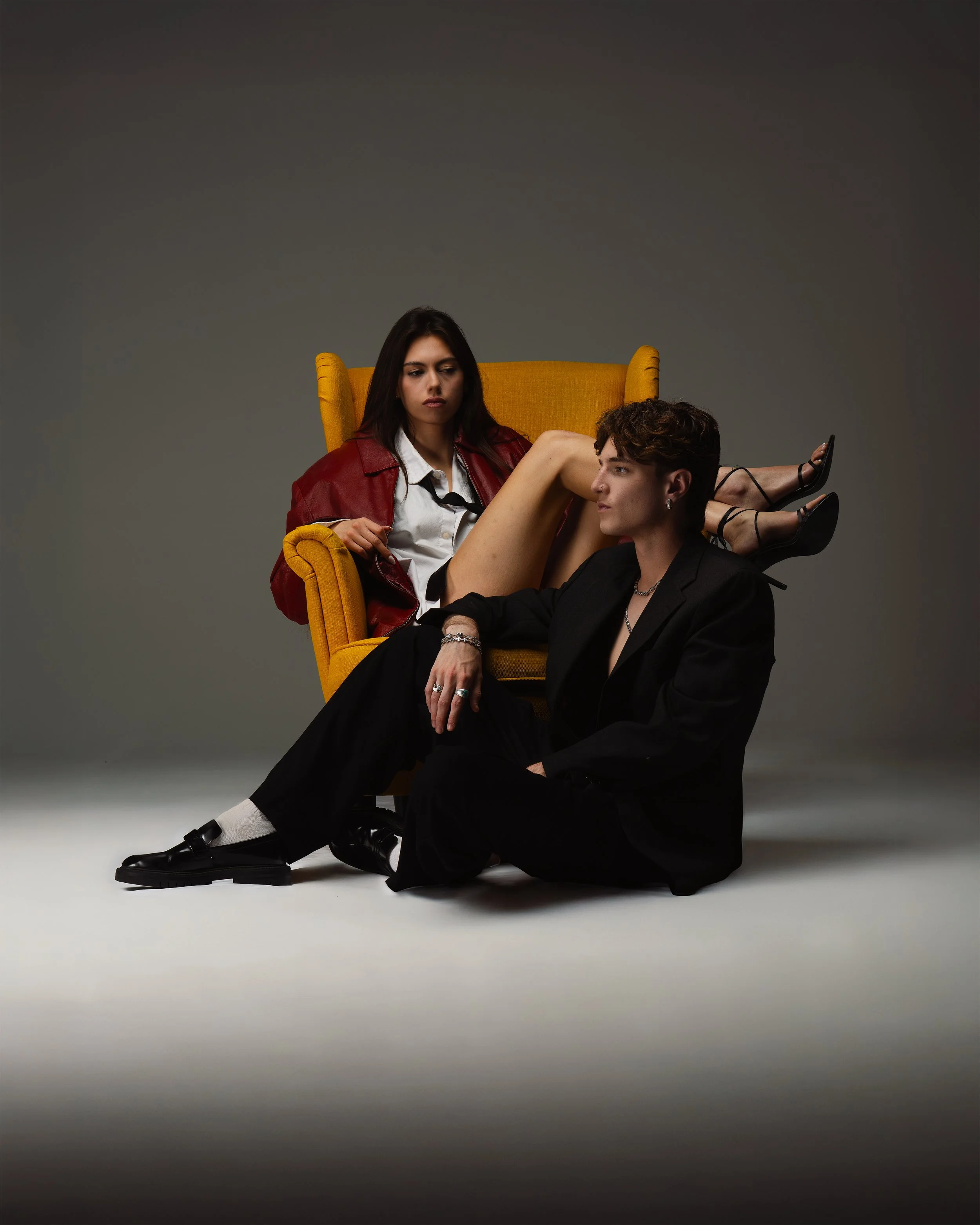 A woman sitting on a yellow armchair with her legs over a man sitting on the floor in front of her, in a studio setting with a gray background.