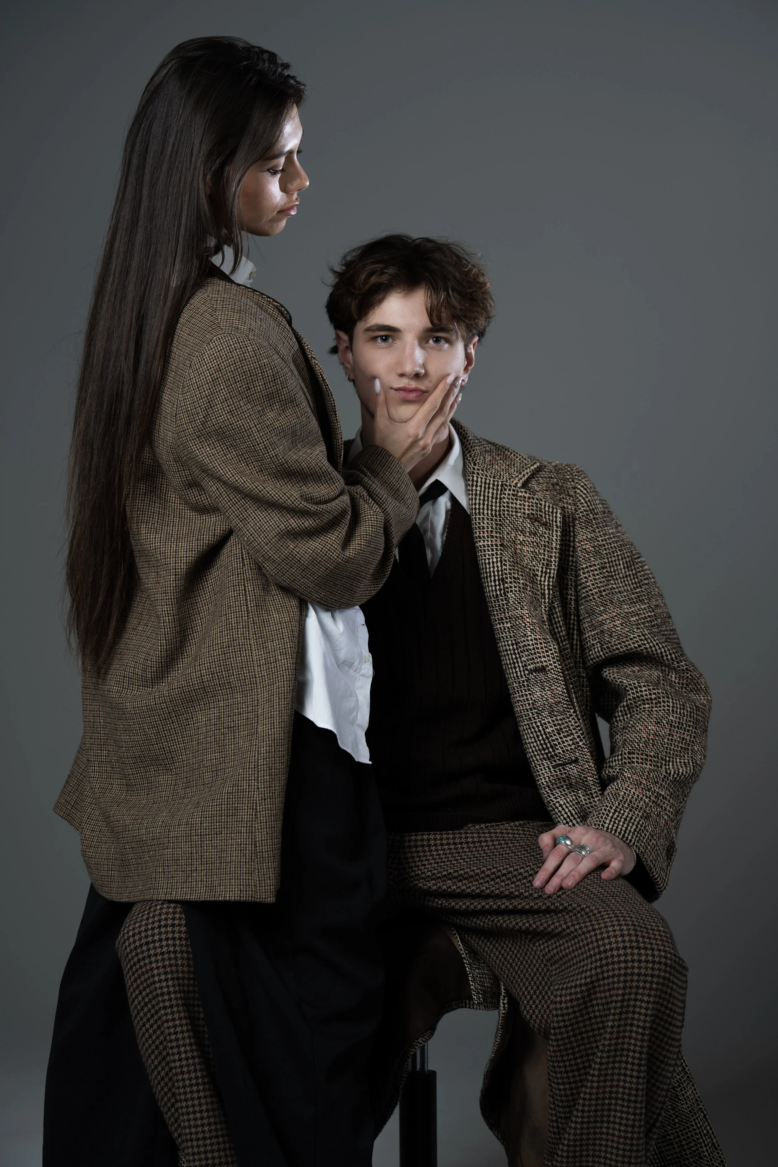 A woman with long brown hair gently touching the face of a young man with curly brown hair, both dressed in brown and checkered suits, against a plain gray background.