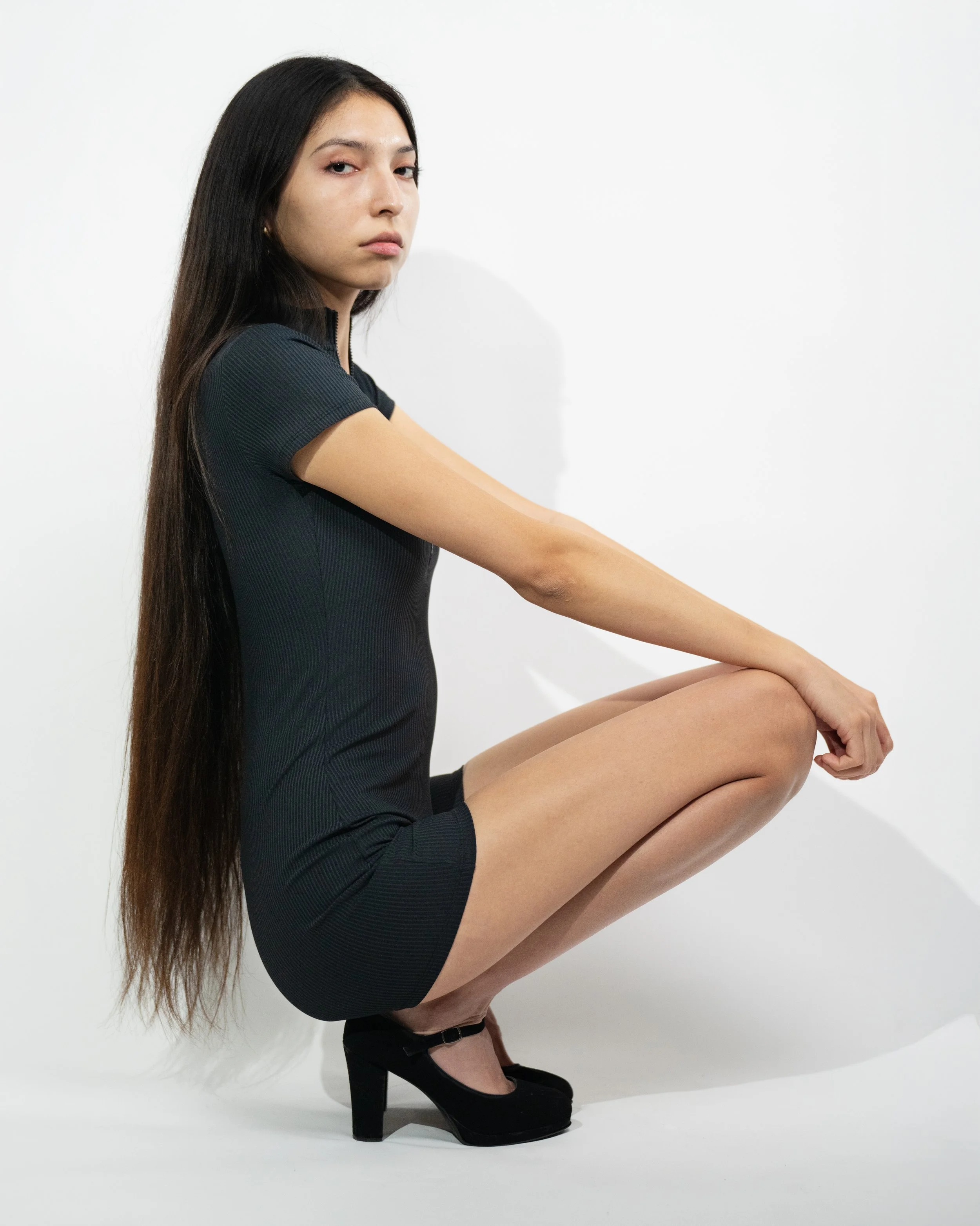 A young woman with long dark hair squatting against a plain white background, wearing a black dress and black high-heeled shoes.