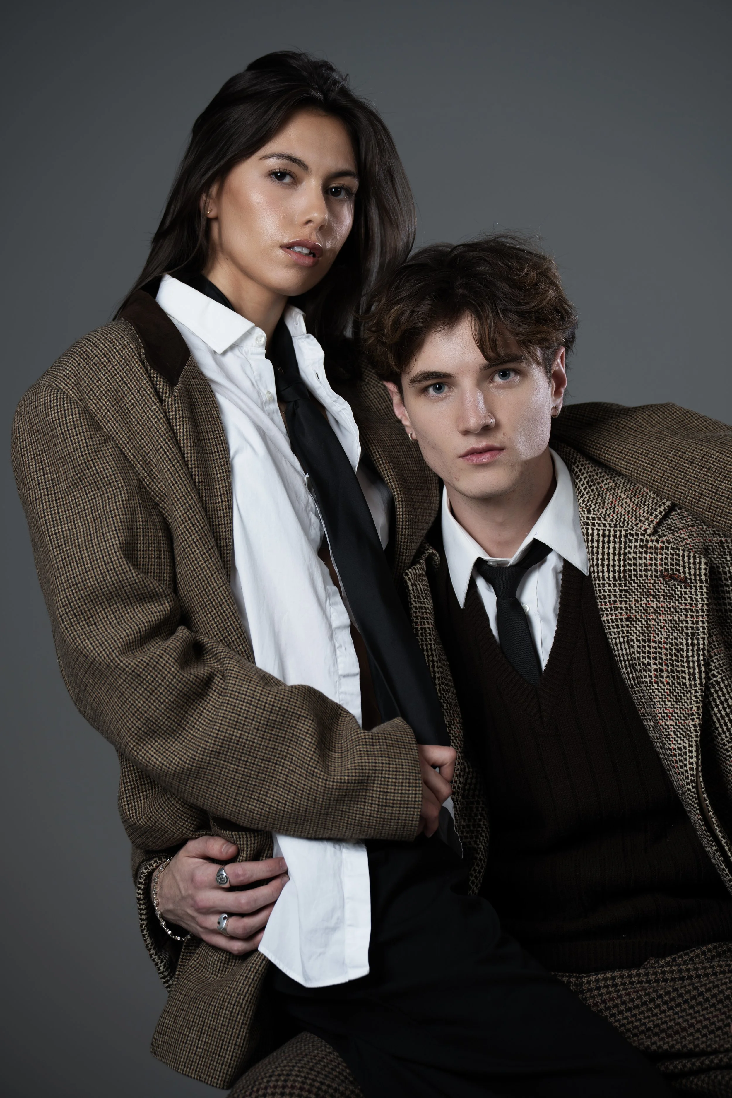 A woman and a young man dressed in vintage-style business attire against a plain gray background.