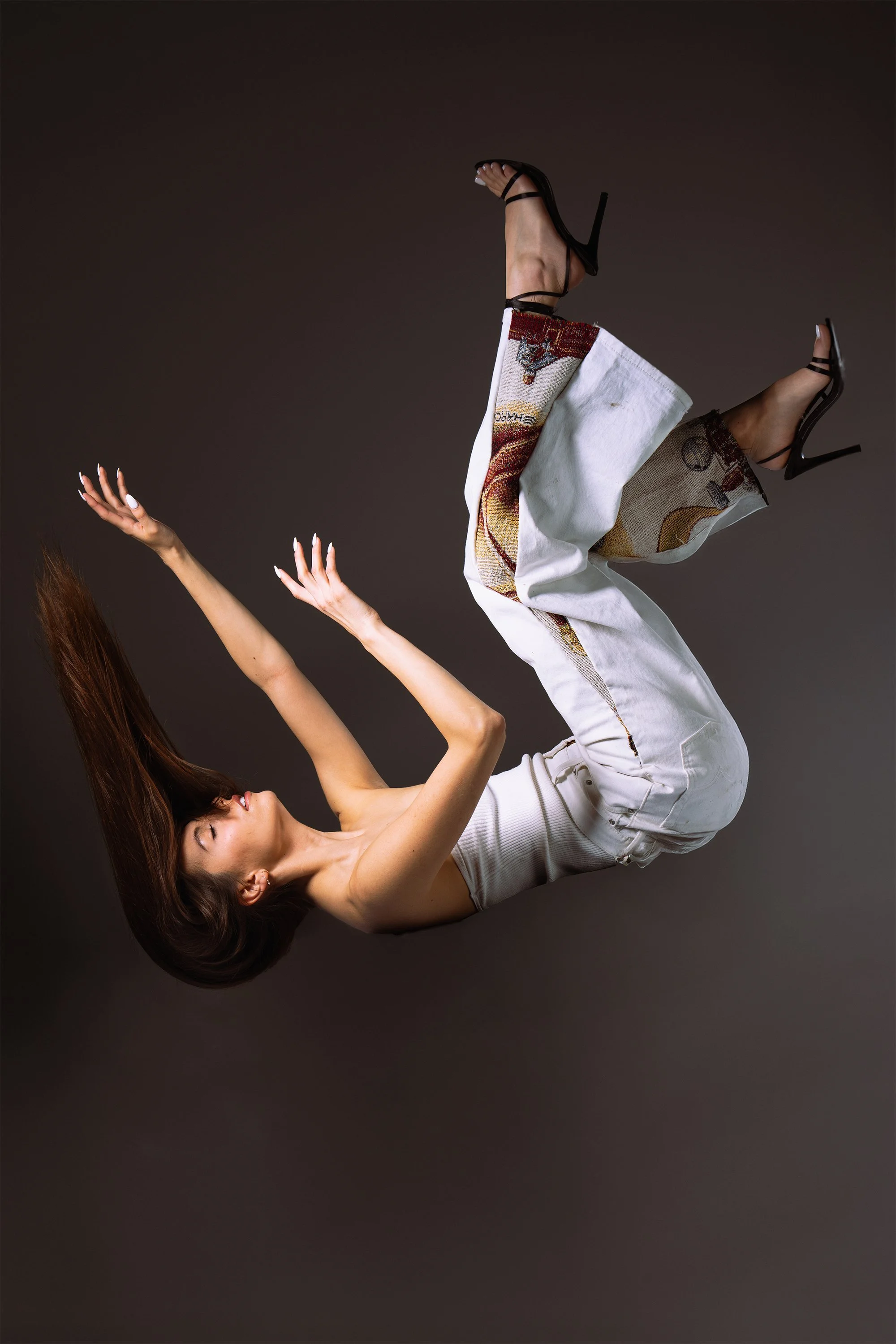 A woman with long hair in a white sleeveless top and embroidered pants, wearing high-heeled black sandals, appears to be floating or falling against a dark background.