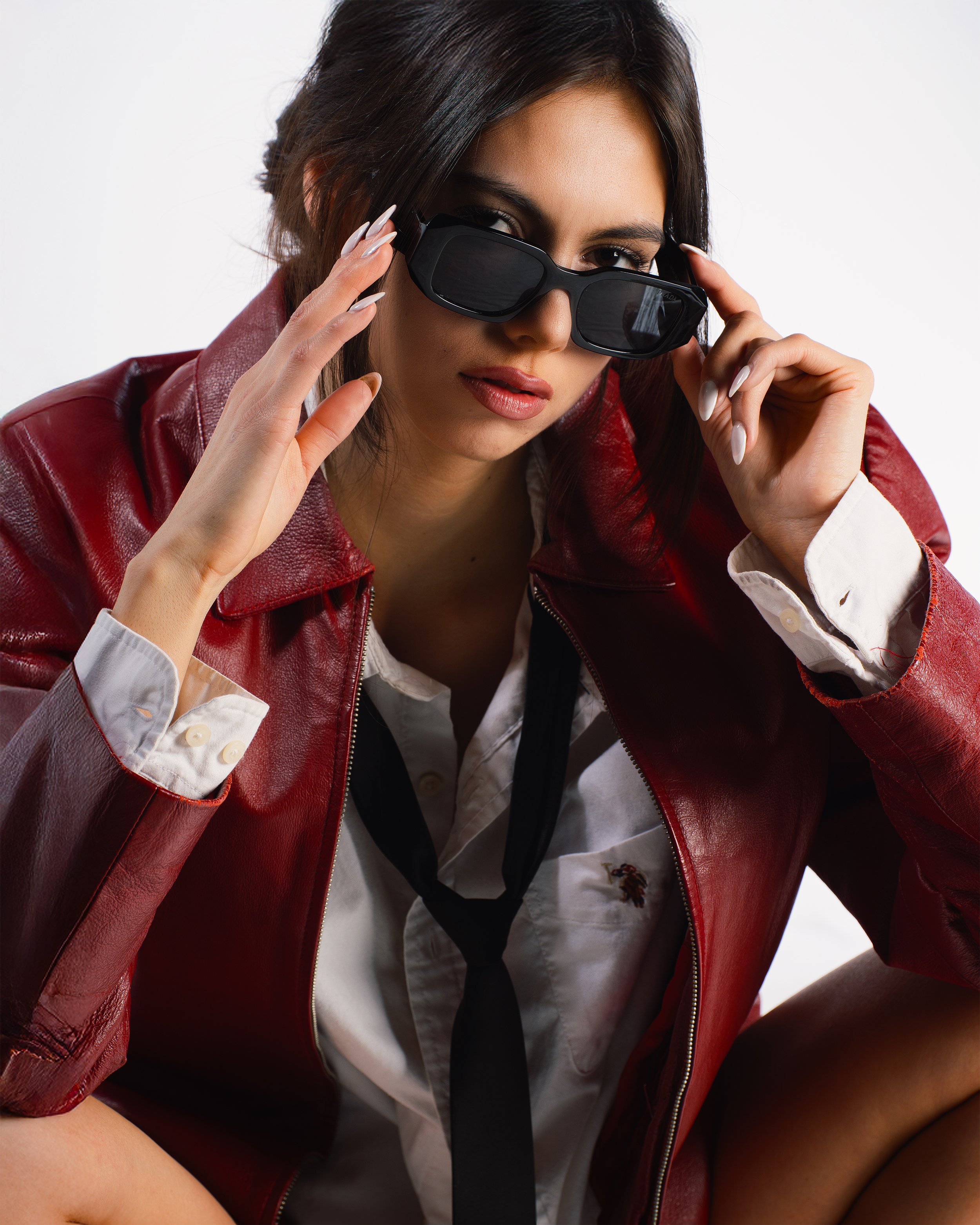 A woman wearing a red leather jacket, sunglasses, a white shirt, and a black tie, looking at the camera.