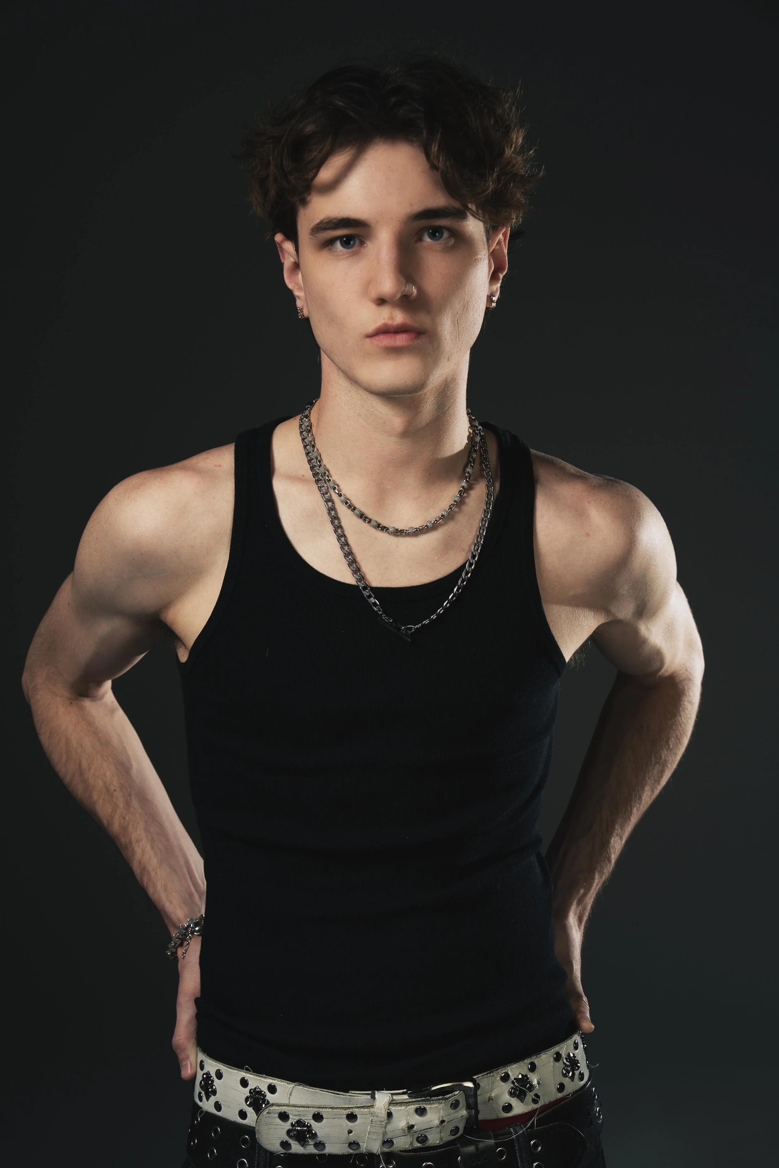 Young man with dark curly hair and earrings, wearing a black tank top and layered silver necklaces, standing with hands on hips against a dark background.