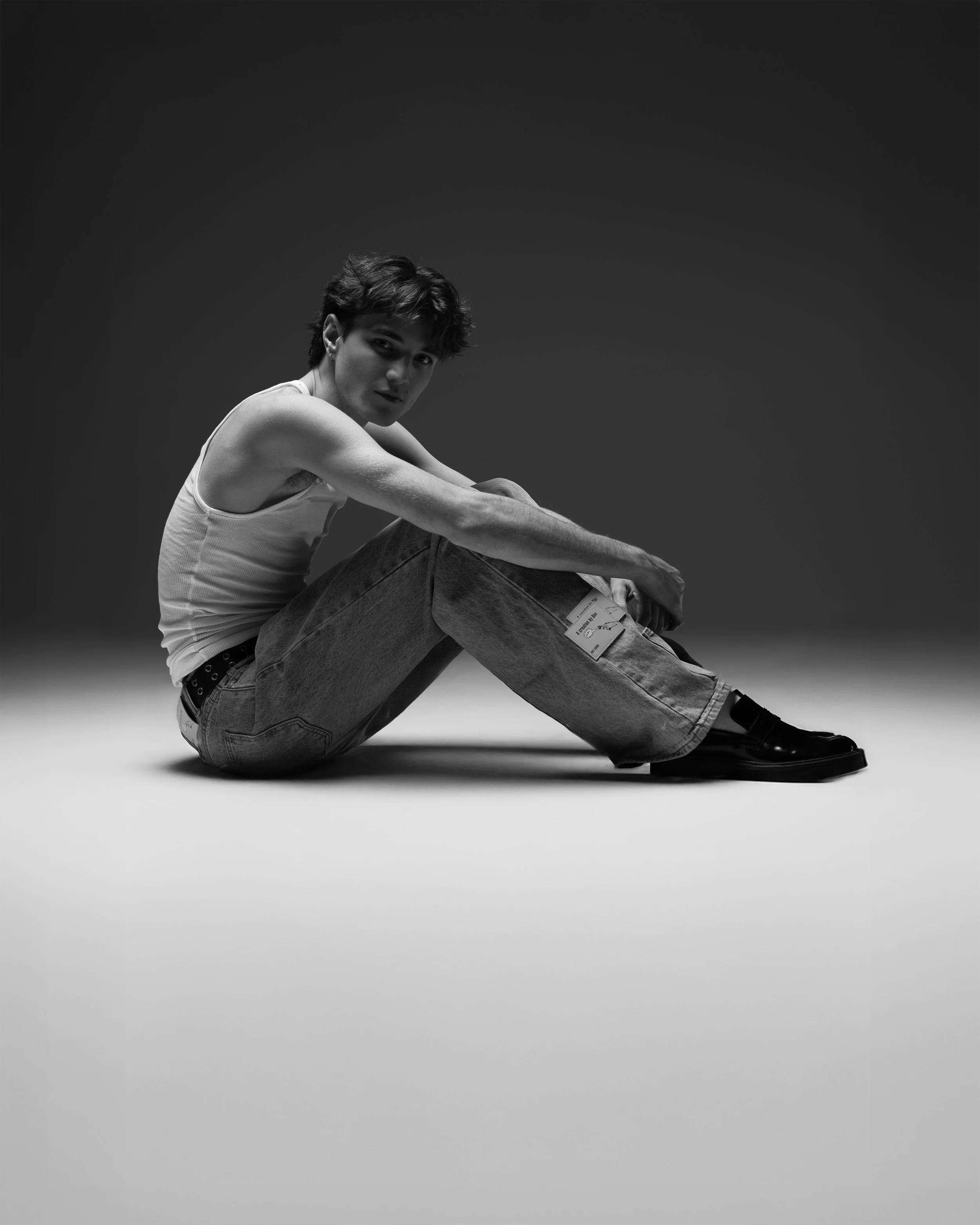 Black and white photo of a young man sitting on the floor with his legs crossed, wearing a tank top, jeans, and shoes, looking into the camera.