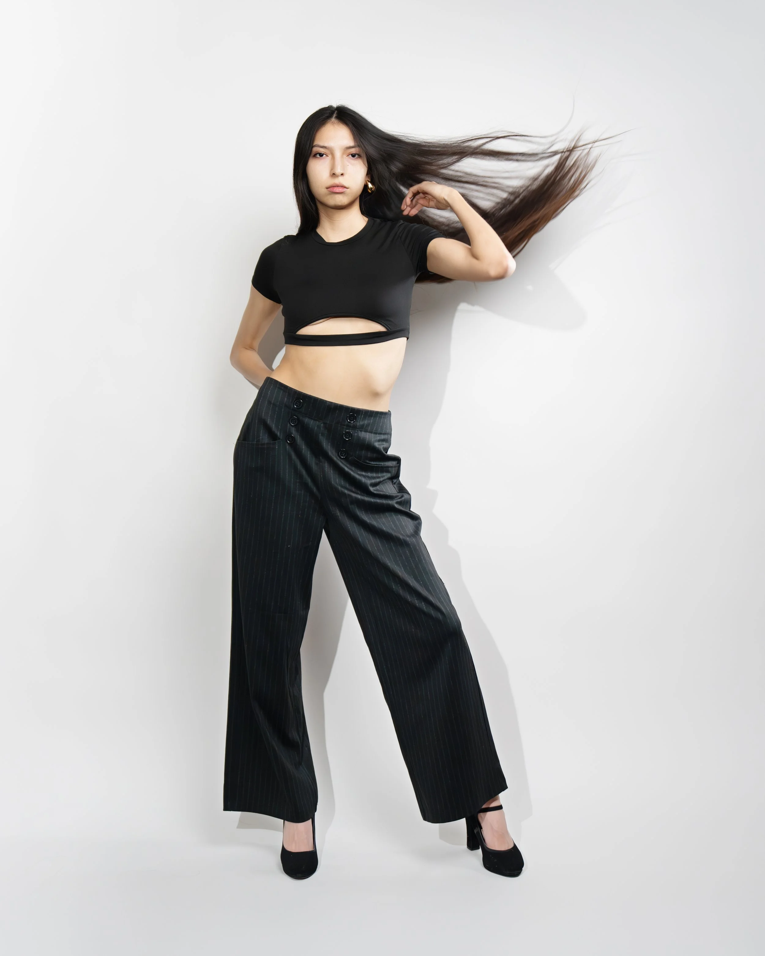 A woman with long dark hair wearing a black crop top and black pinstripe pants posing against a plain white background.