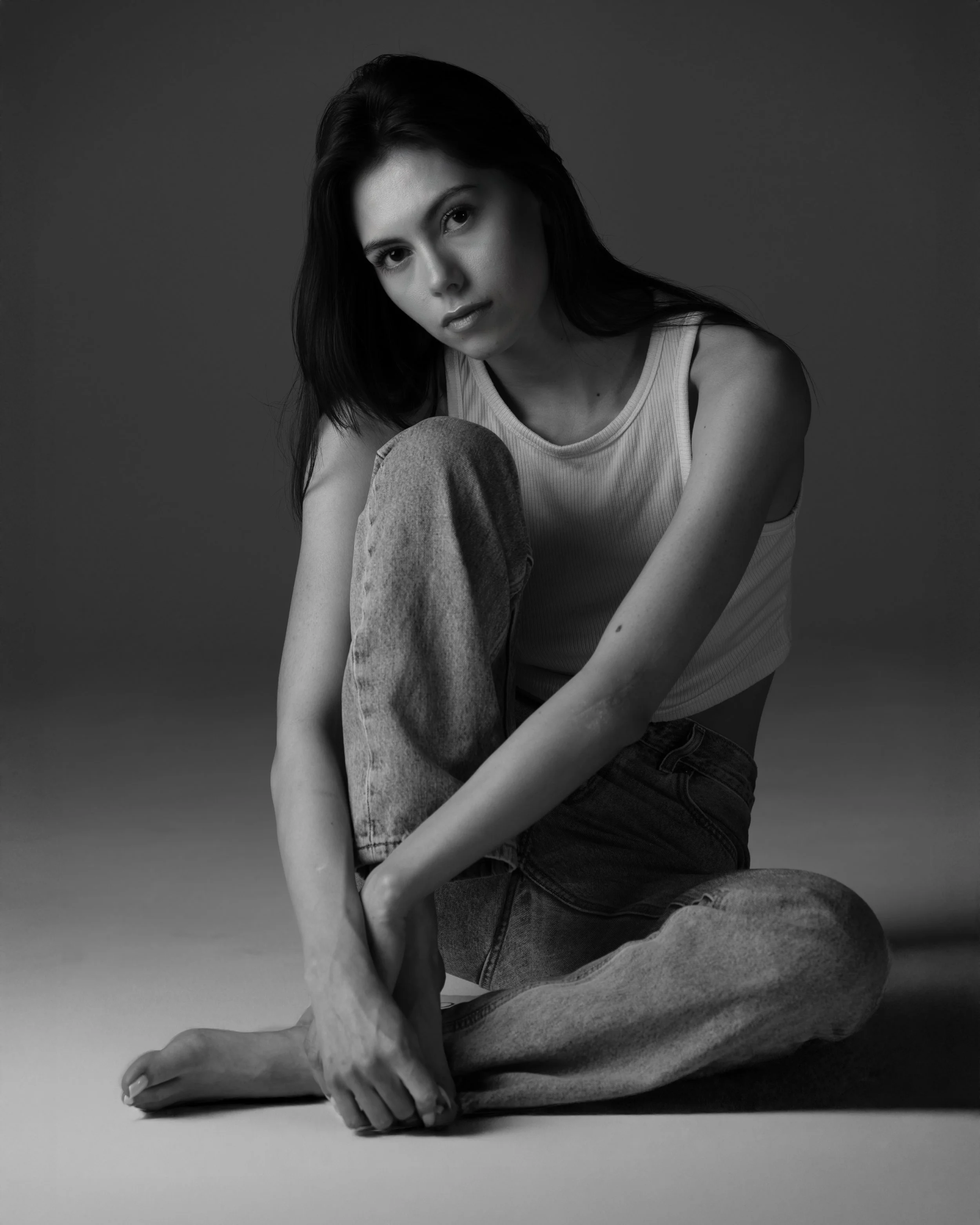 A woman with dark hair sitting on the floor, wearing a sleeveless shirt and jeans, looking at the camera in a black and white photograph.