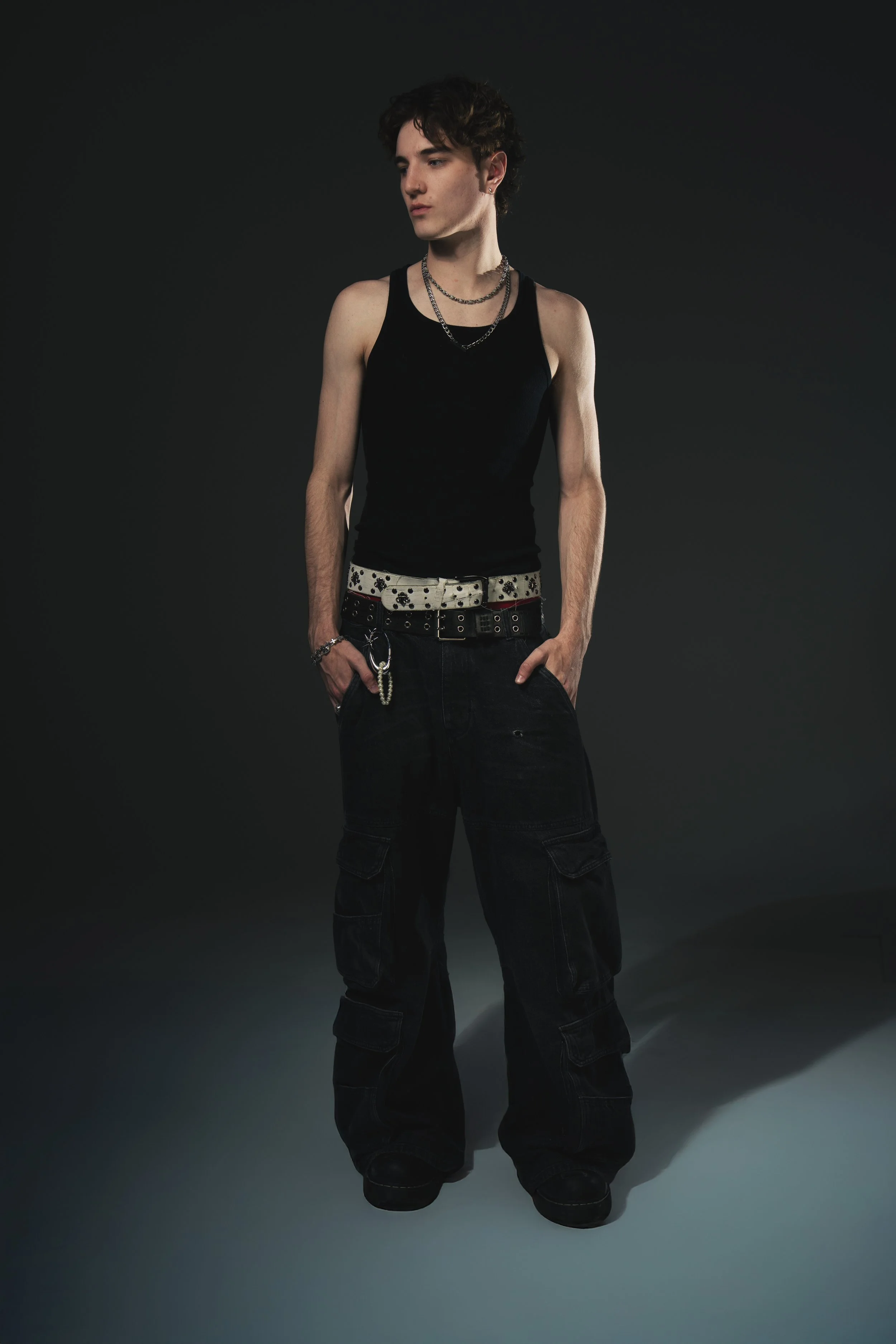 A young man with curly dark hair wearing a black tank top, layered necklaces, and black cargo pants standing against a dark background.