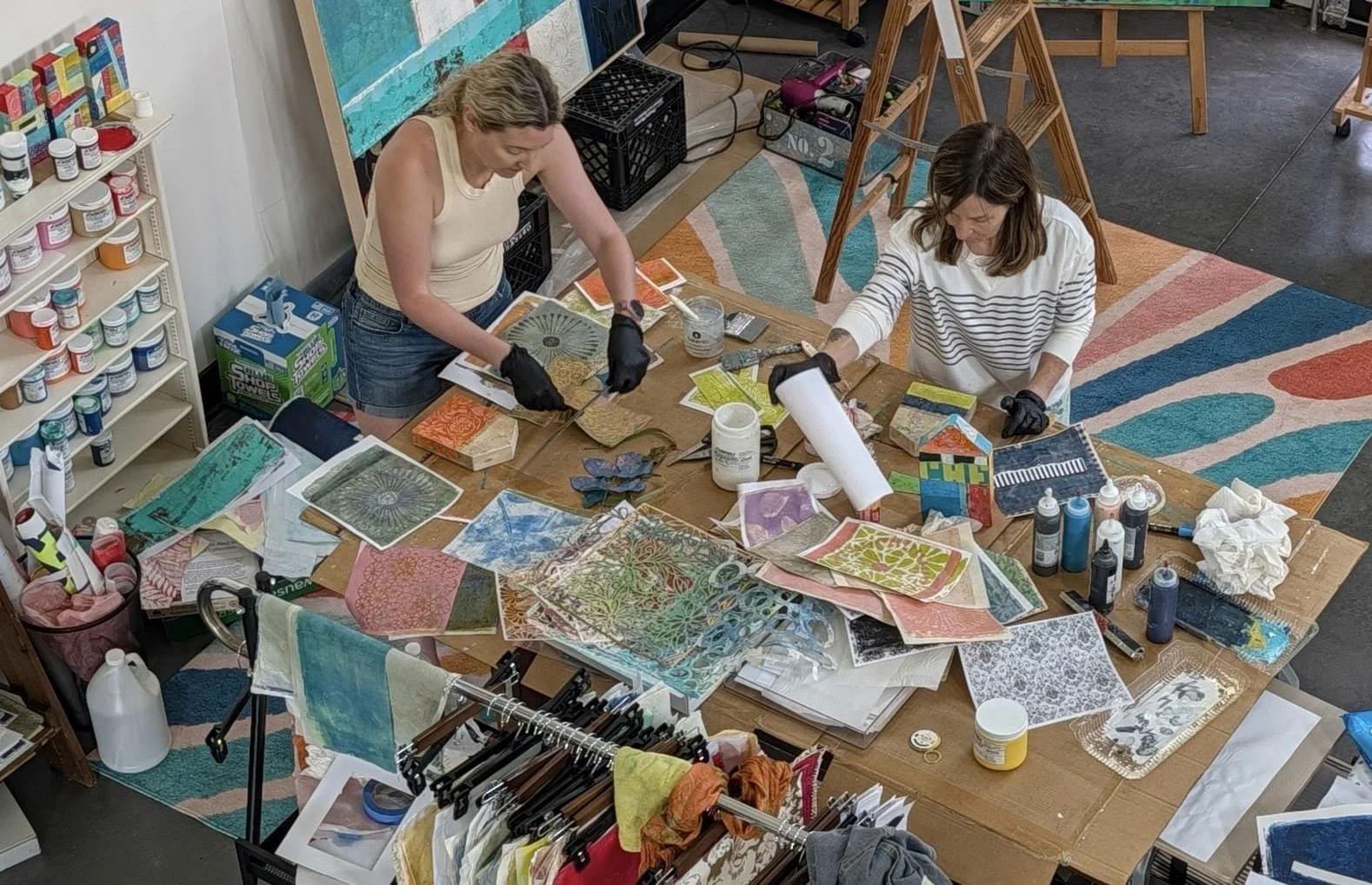 Two women working on art projects at a cluttered table with paint bottles, papers, fabric, and tools in an art studio.