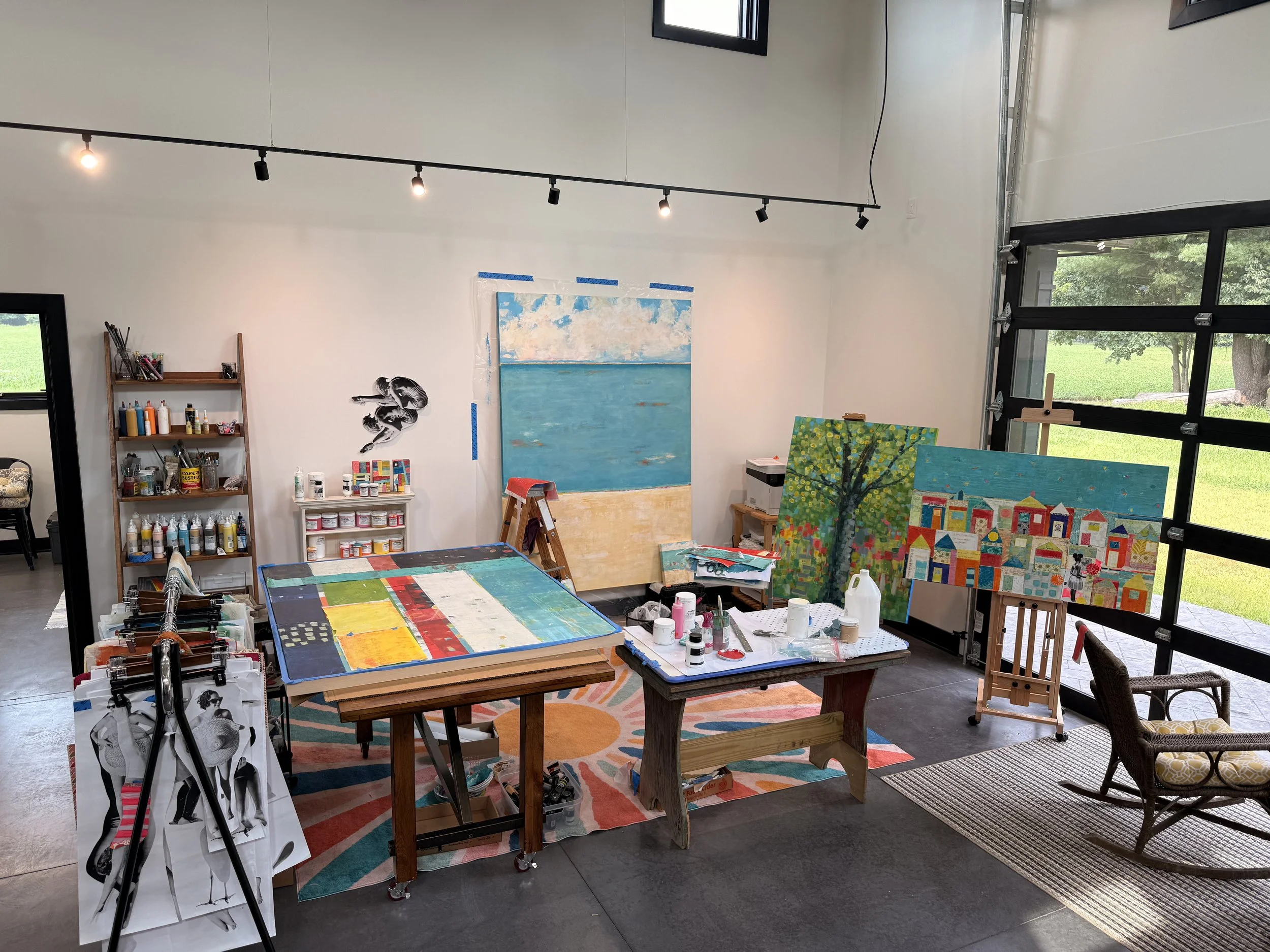 An art studio with paintings on the wall, art supplies on a shelf, and multiple paintings in progress. Large windows allow natural light into the space.