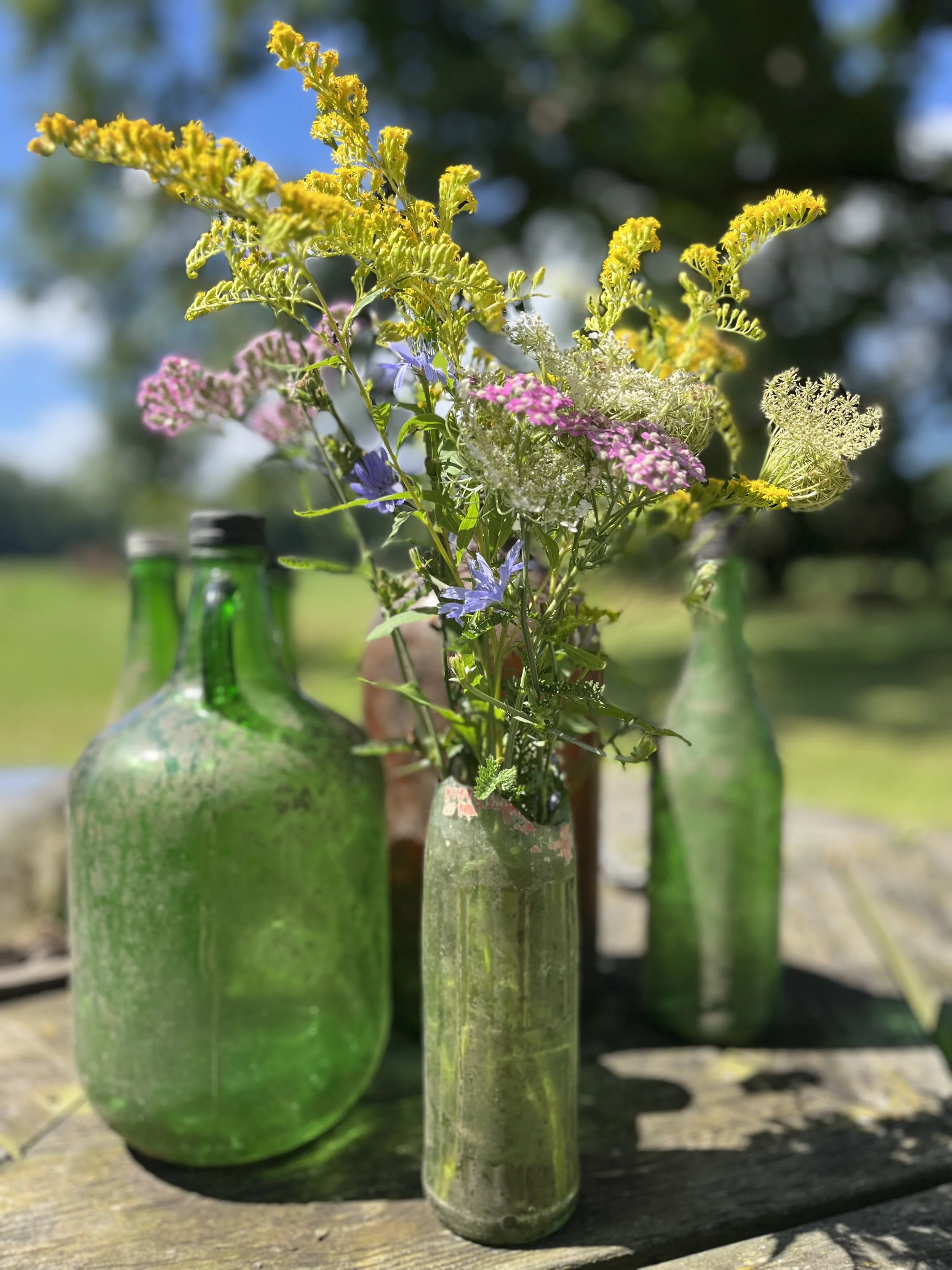 Three glass bottles with flowers arranged inside sitting on a wooden surface outdoors, with a blurred background of trees and sky.