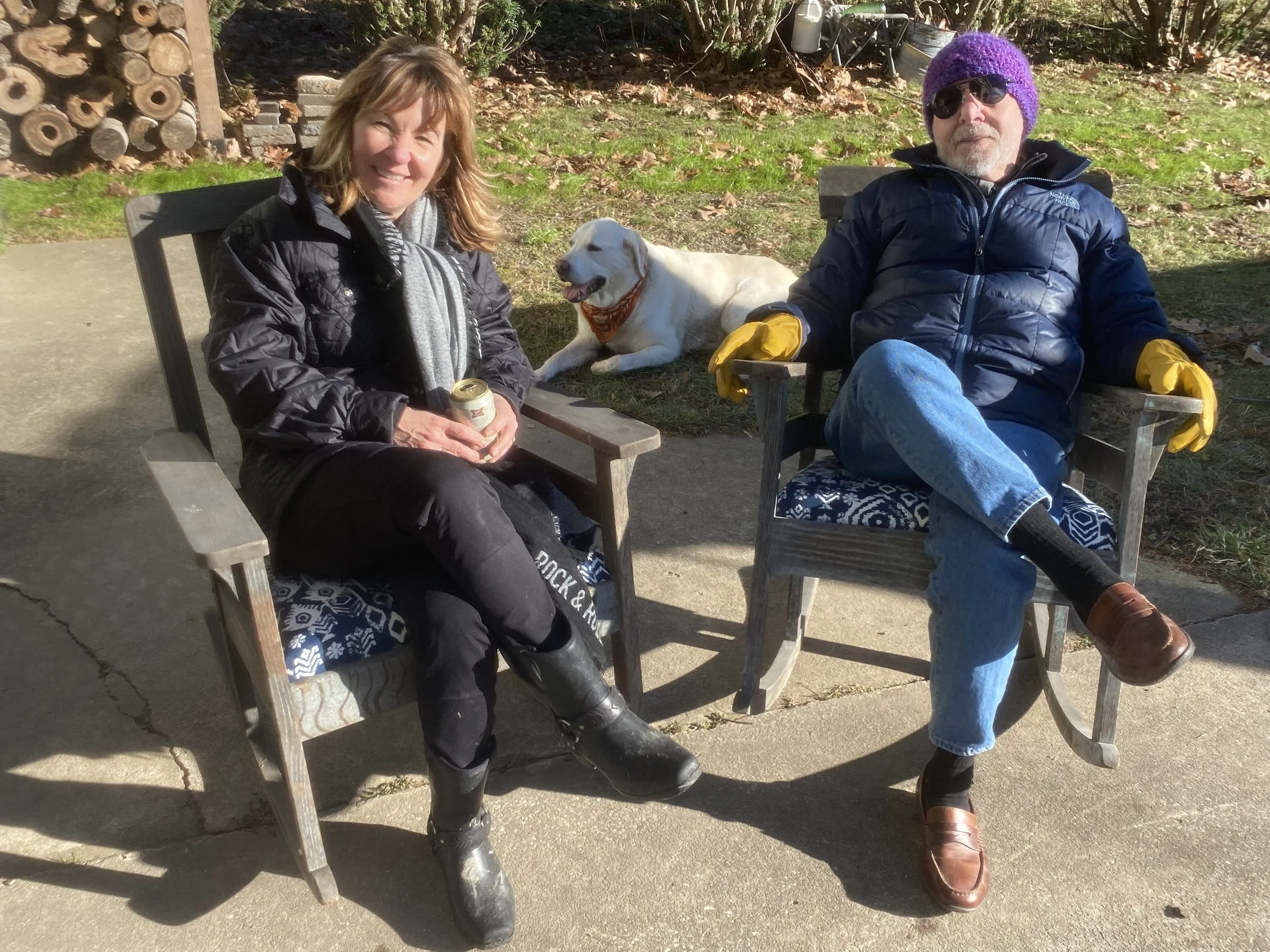 Two people sitting outdoors on benches, smiling at the camera with a white Labrador dog lying between them. The woman on the left has light brown hair, wearing a black jacket and black boots, and holding a beverage can. The man on the right has a gra