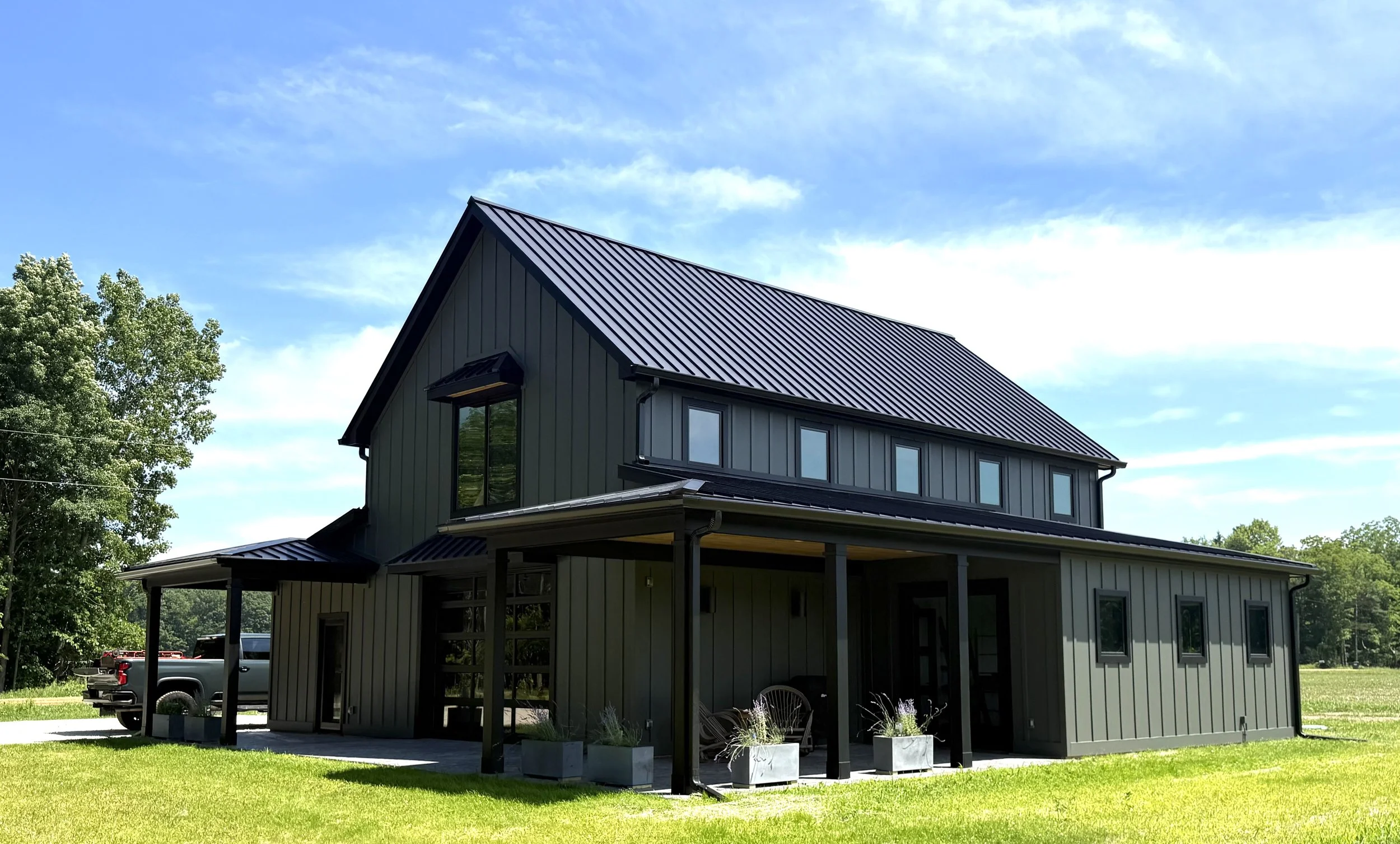 Modern two-story house with metal roof, large windows, gray siding, surrounded by green grass and trees, under a partly cloudy sky.