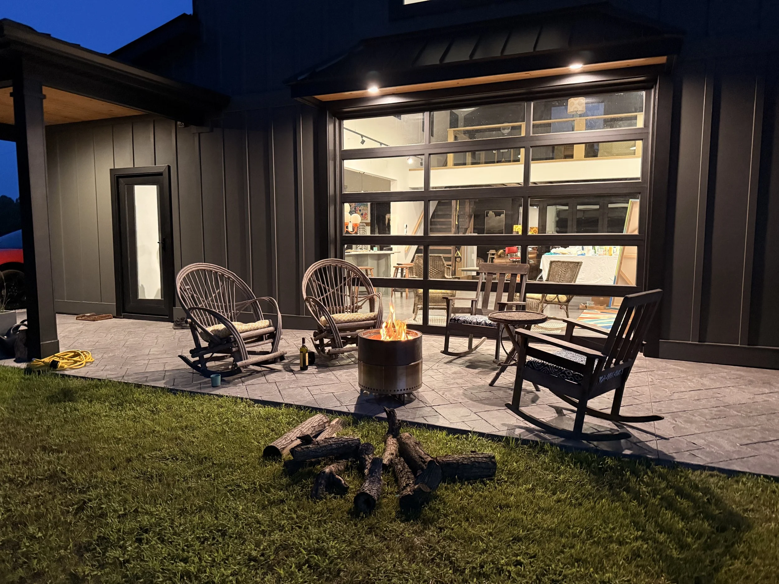 An outdoor patio area at night with a fire pit, wicker chairs, and a modern house with large windows and interior lighting visible.