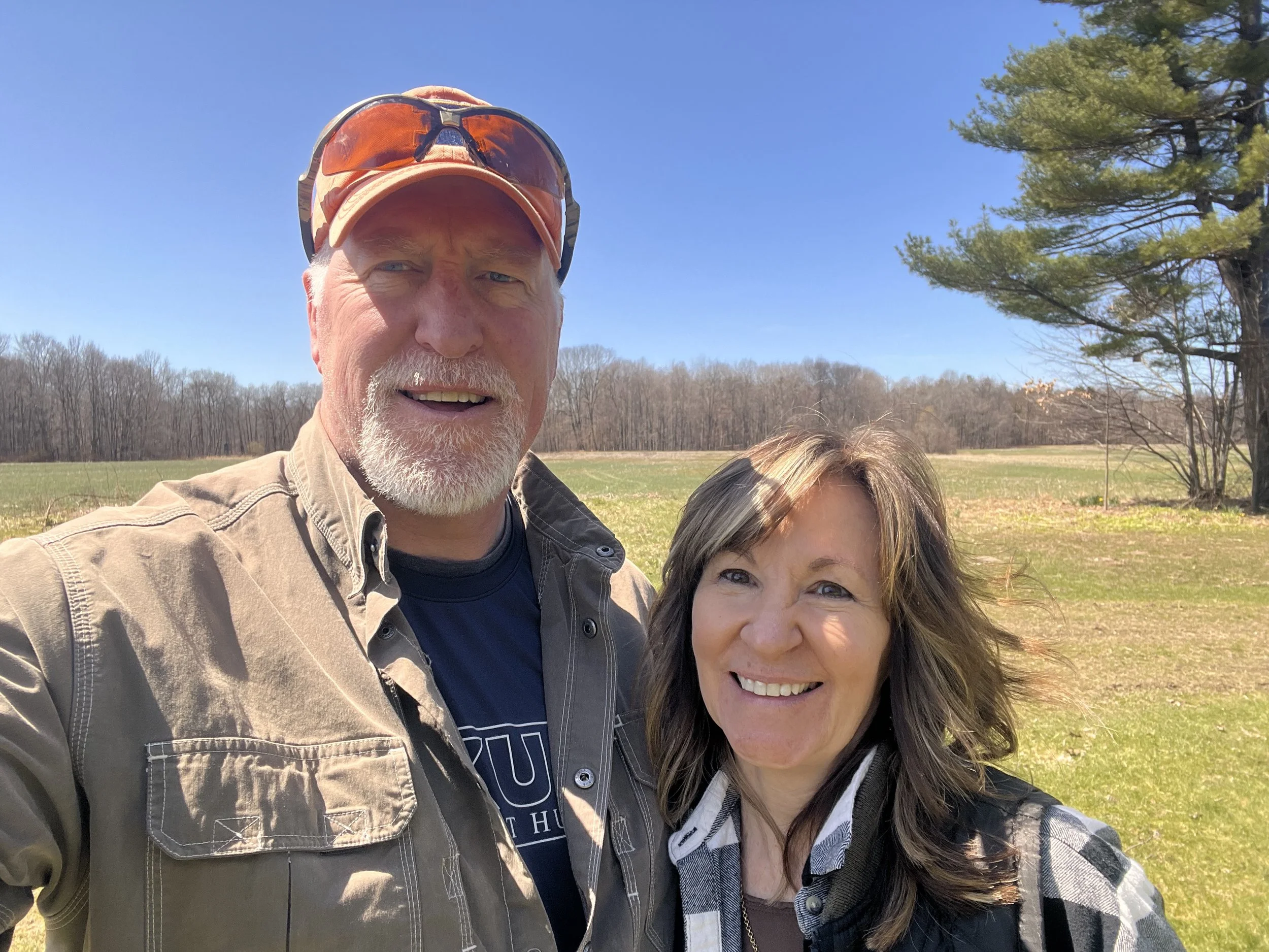 A smiling older man with a white beard and wearing an orange cap with sunglasses on top, standing outdoors alongside a smiling woman with long, dark hair, in a landscape with trees and a blue sky.