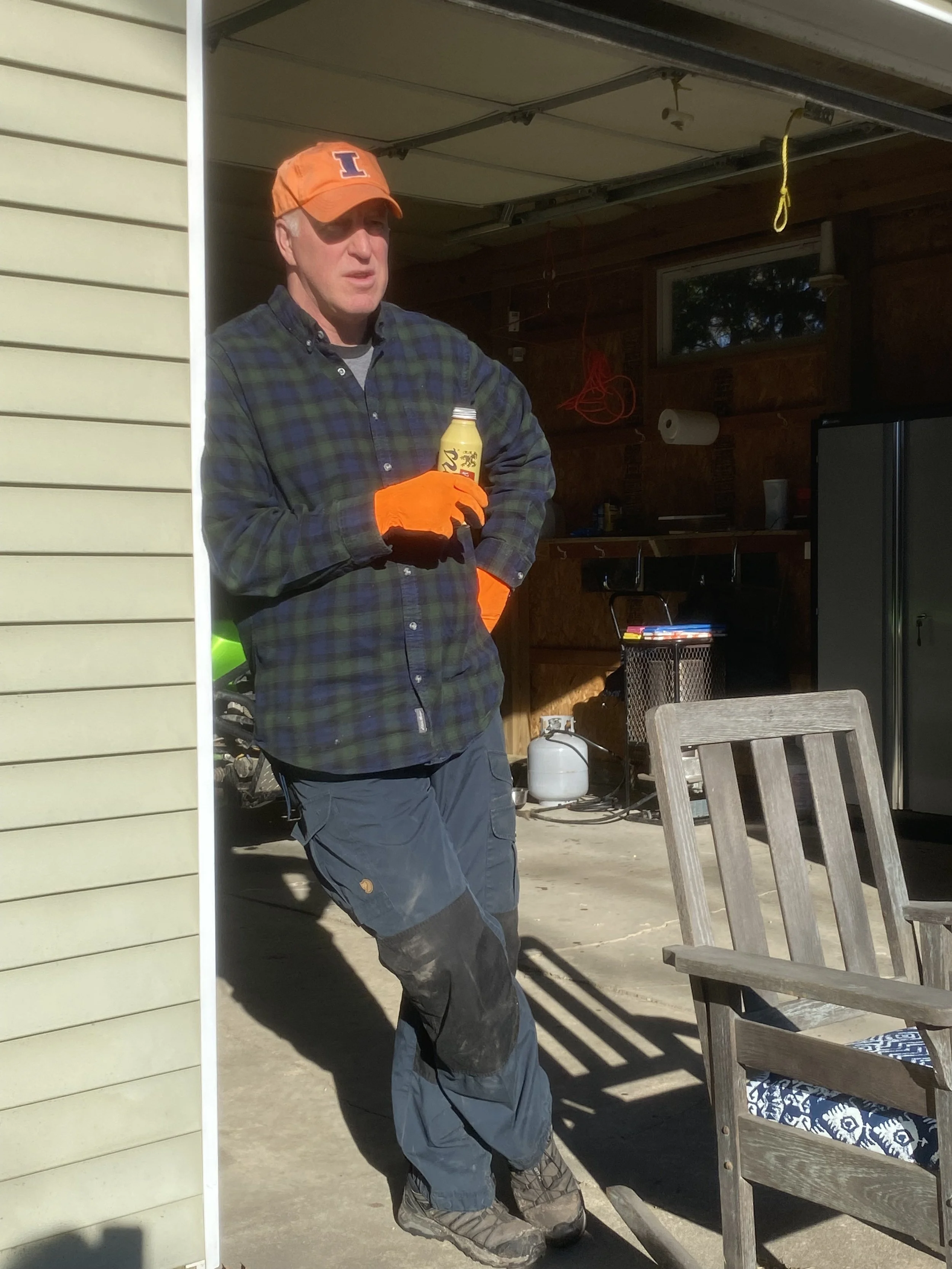 A man wearing an orange cap with a blue letter "I" and bright orange gloves, standing outdoors near a wooden deck, holding a bottle of beer.