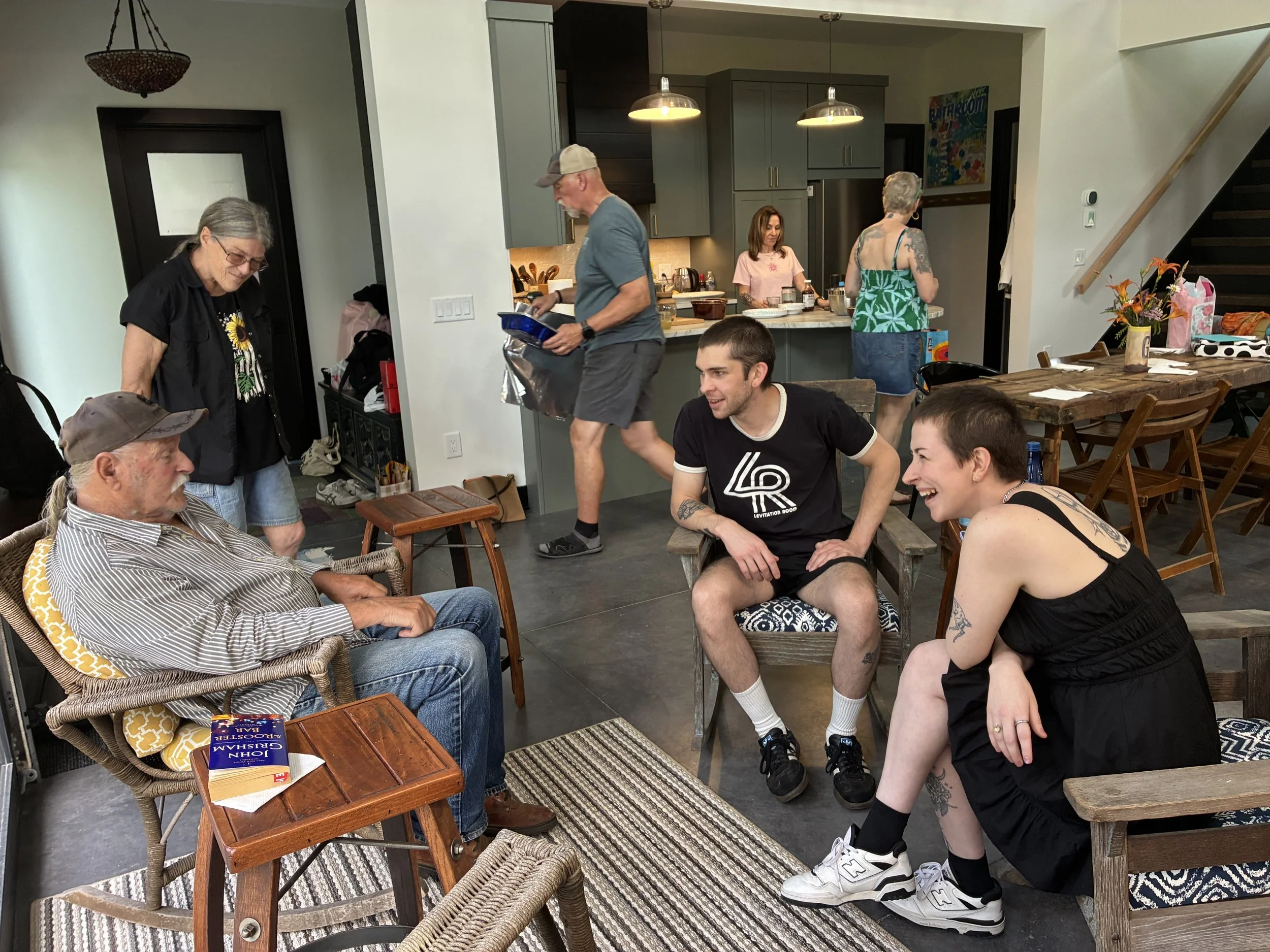 A group of people gathered in a modern kitchen and dining area, engaging in conversation and activities, with a man reading a book and others smiling and talking.