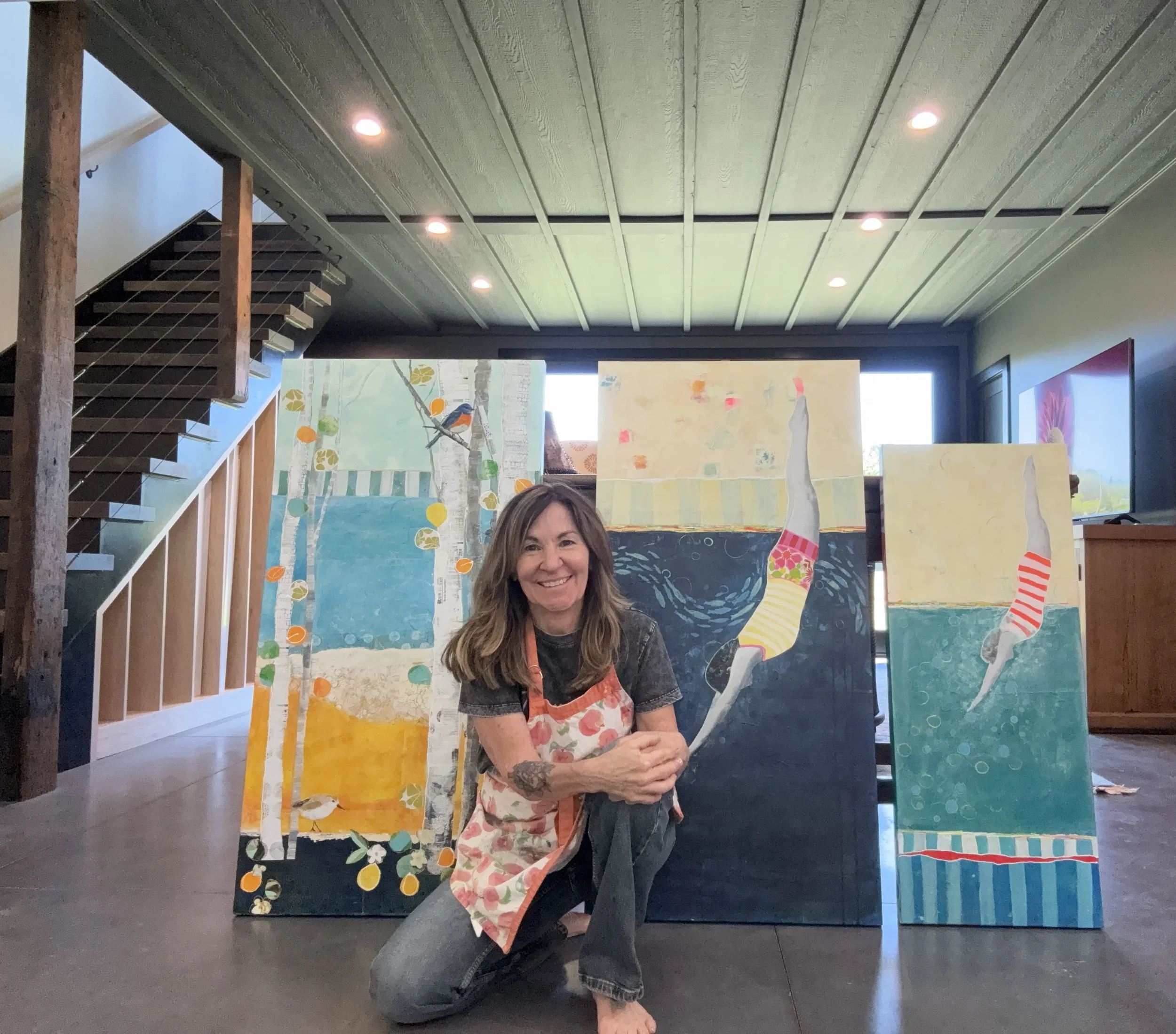 A woman with long hair and tattoos on her arm, wearing a floral apron, smiling and kneeling on one knee inside an art gallery with several paintings on display and a staircase on the left side.