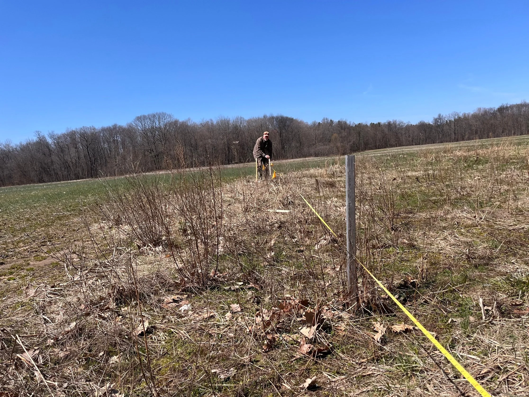 A man in a gray jacket and sunglasses standing in a field with dry grass and plants, holding a yellow flag or ribbon, with a background of leafless trees under a clear blue sky.