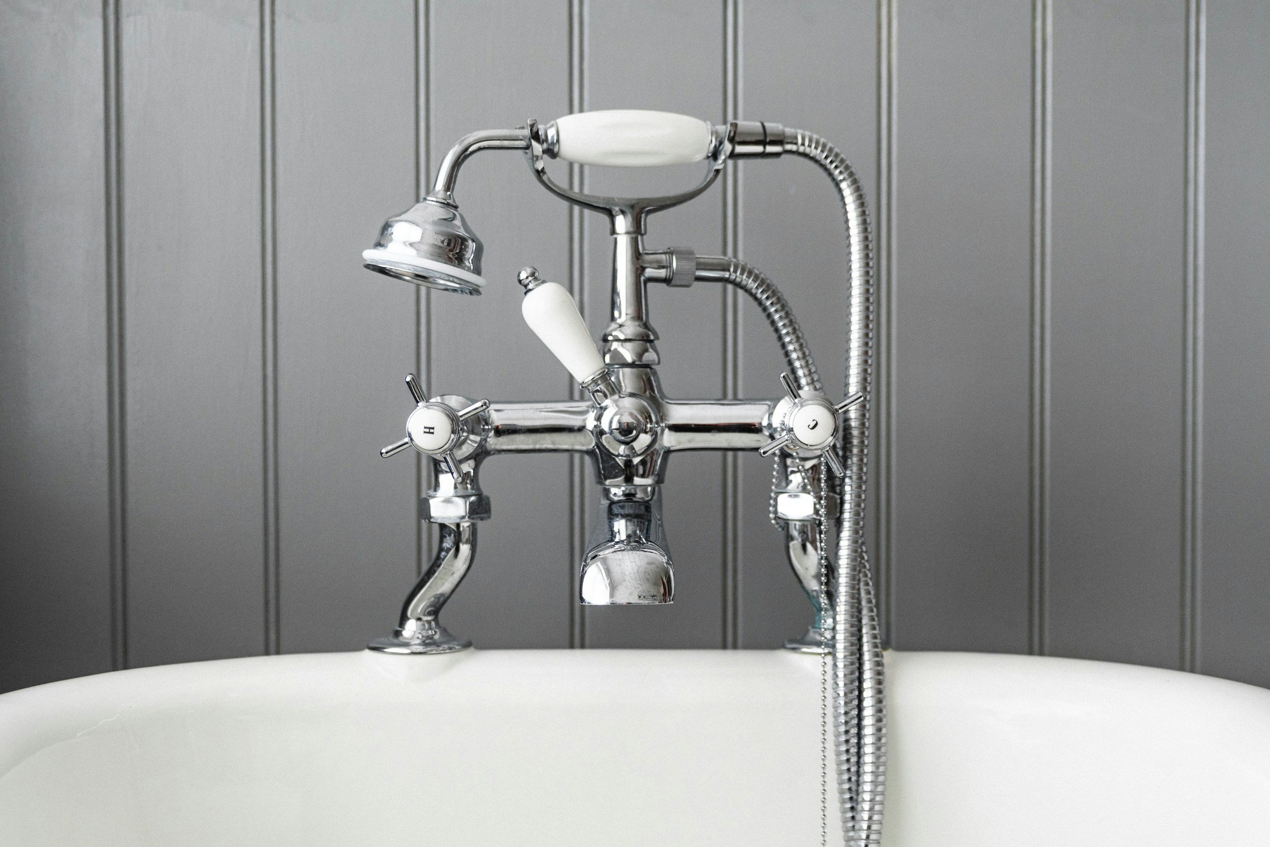Close-up of a vintage-style silver bathtub faucet with separate hot and cold water knobs, a handheld showerhead, and a gray paneled wall background.