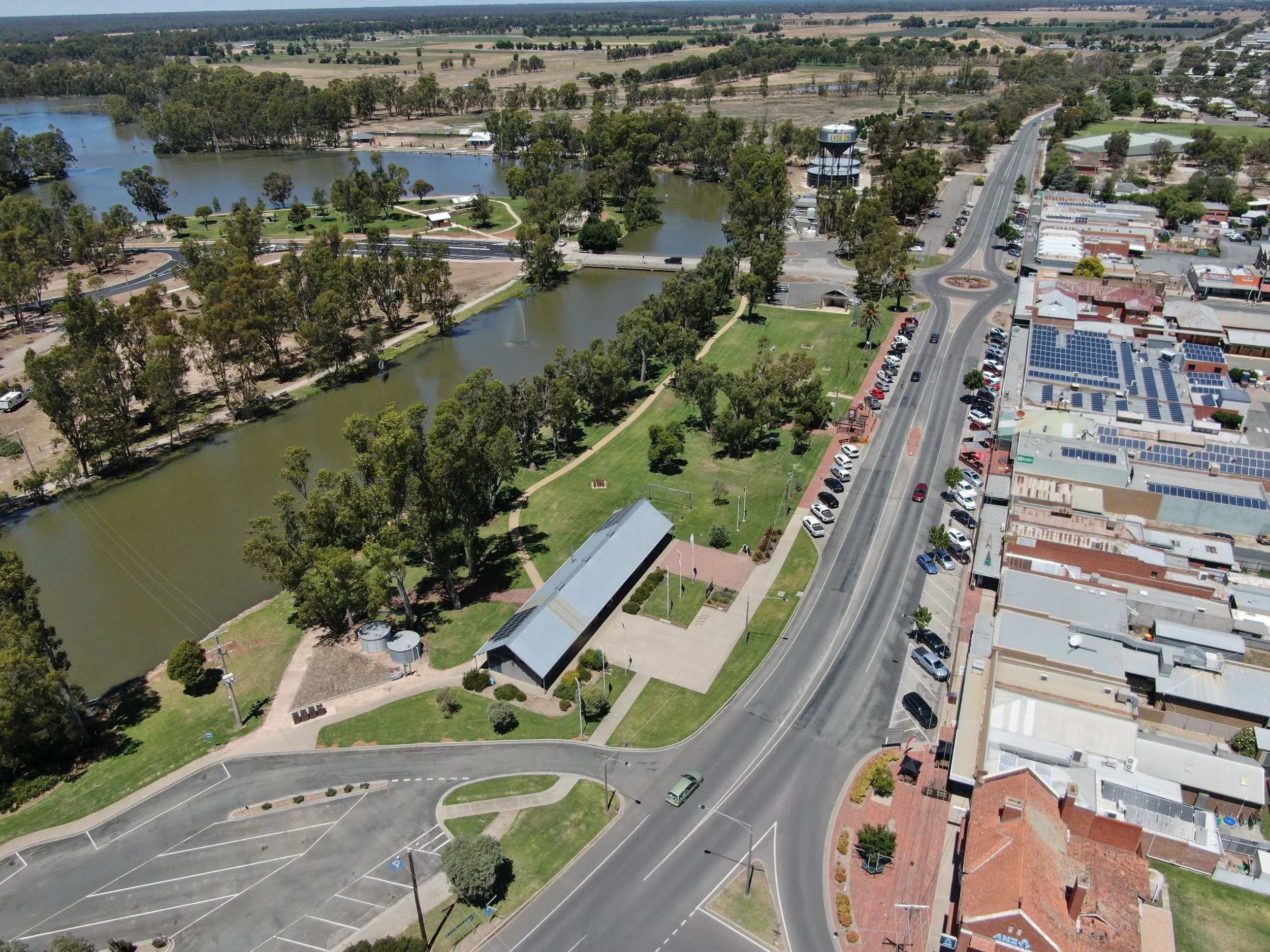 An aerial view of a small town park with water features and surrounding urban area. The park has lakes, walking paths, trees, picnic areas, and a pavilion. Adjacent to the park is a main street with parked cars, shops, and businesses, with a water tower visible in the background.