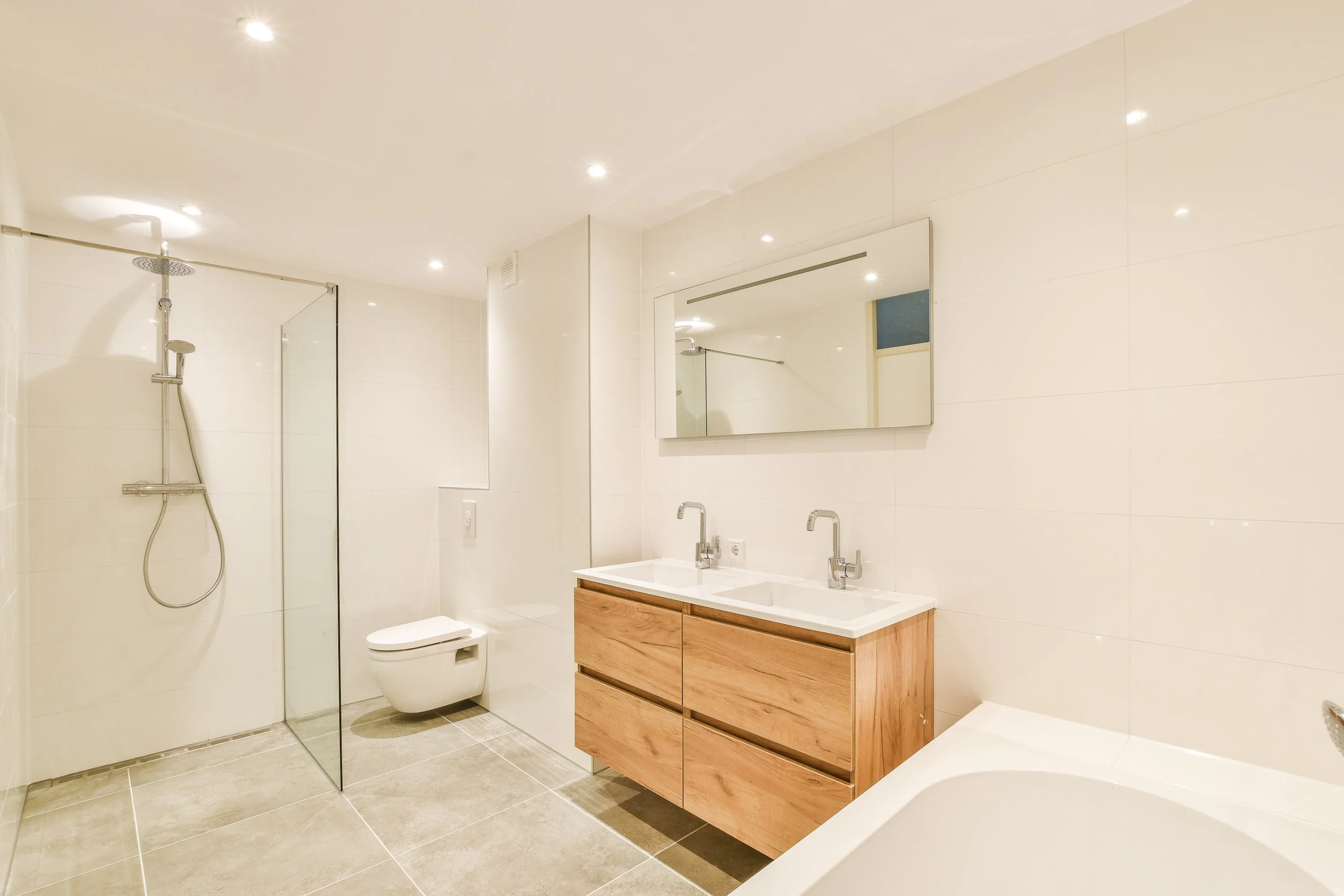 Modern bathroom with walk-in shower, wall-mounted toilet, double sink vanity with wooden cabinets, large mirror, bathtub, and recessed ceiling lights.