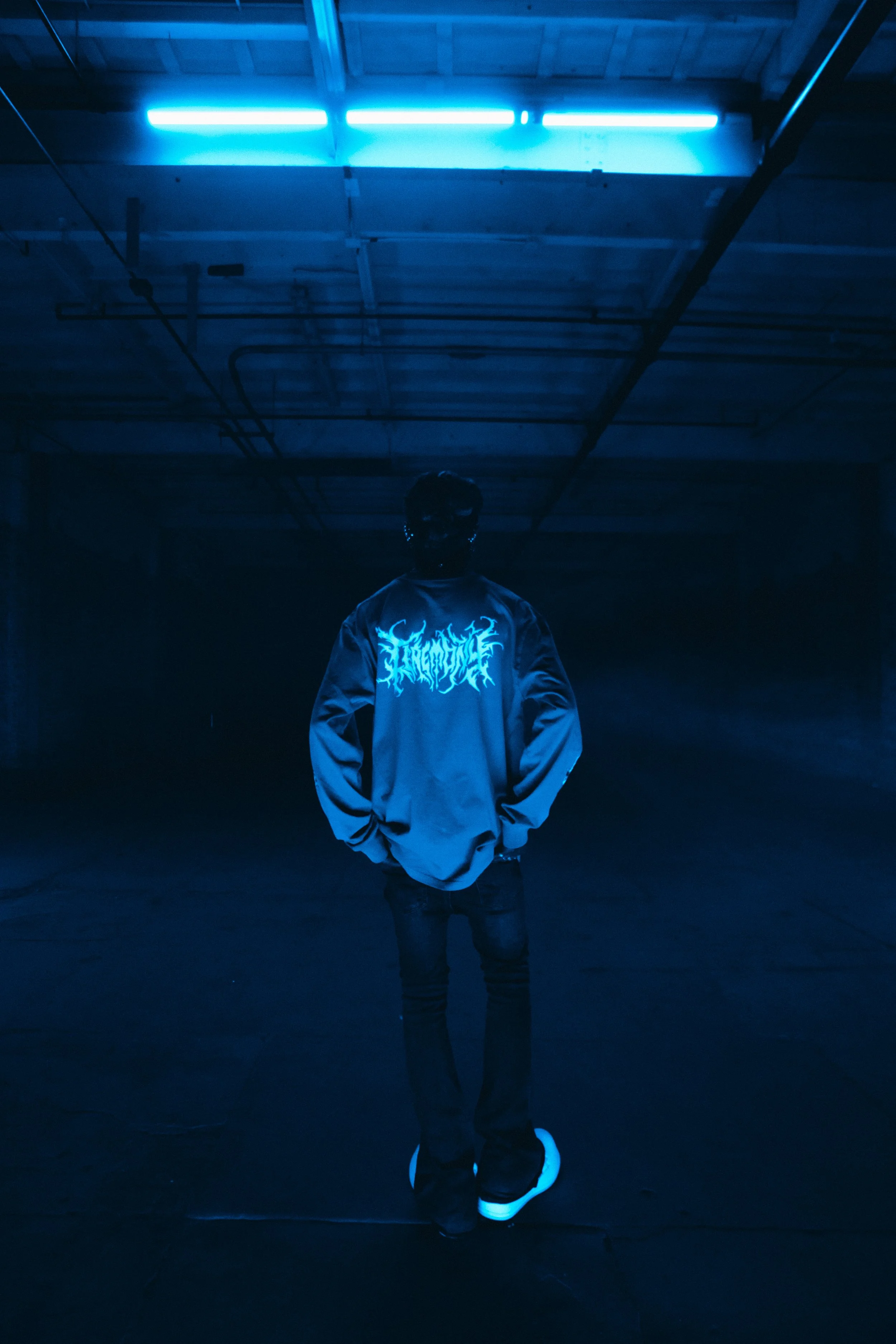Person in a blue-lit room wearing a black cap and jacket with white Ceremony text on the back, standing under a blue fluorescent light.