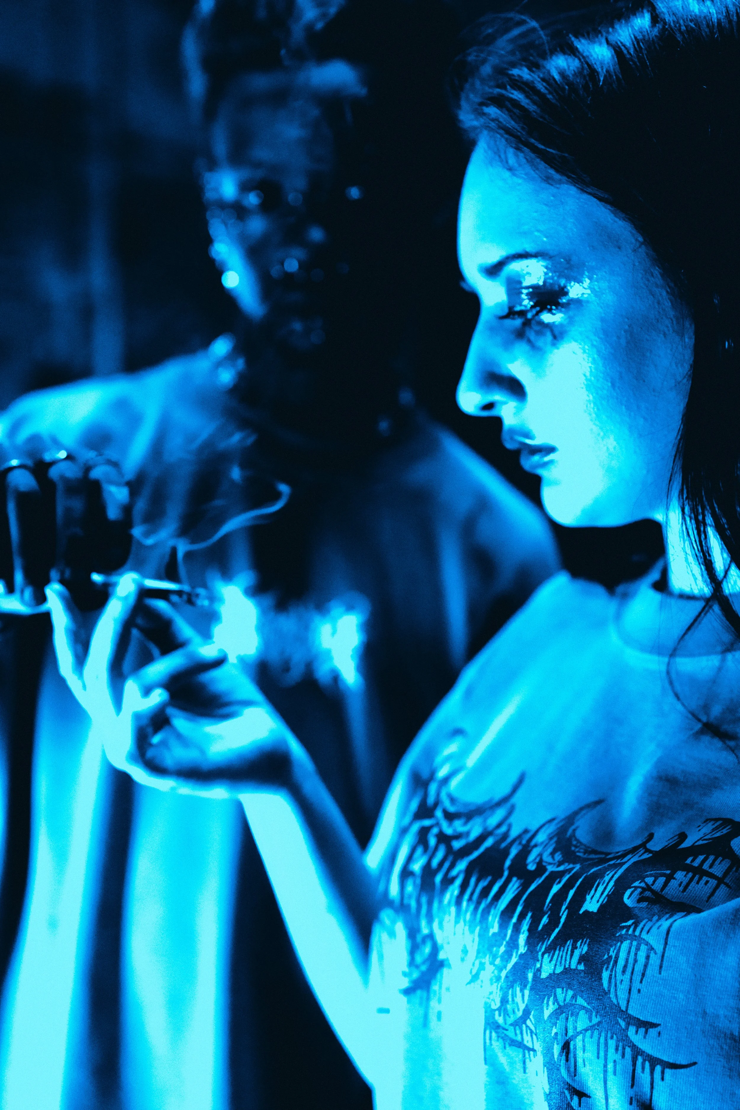 Two people in a dark room under blue lighting; one is holding a lit Ceremony preroll, and the other has glitter on their face.
