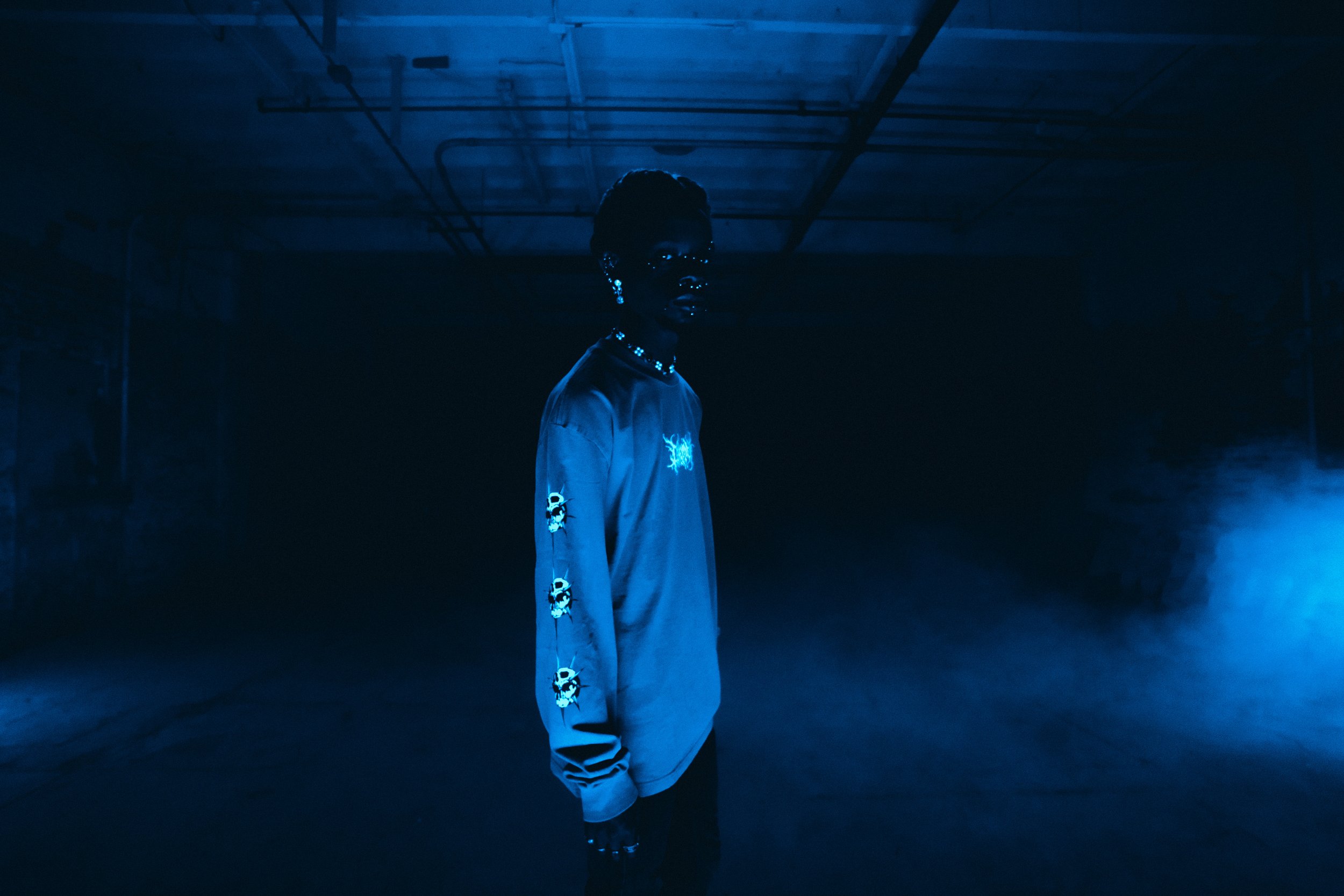 Person in dark room with blue lighting wearing a long-sleeve Ceremony shirt with skull designs, surrounded by mist.