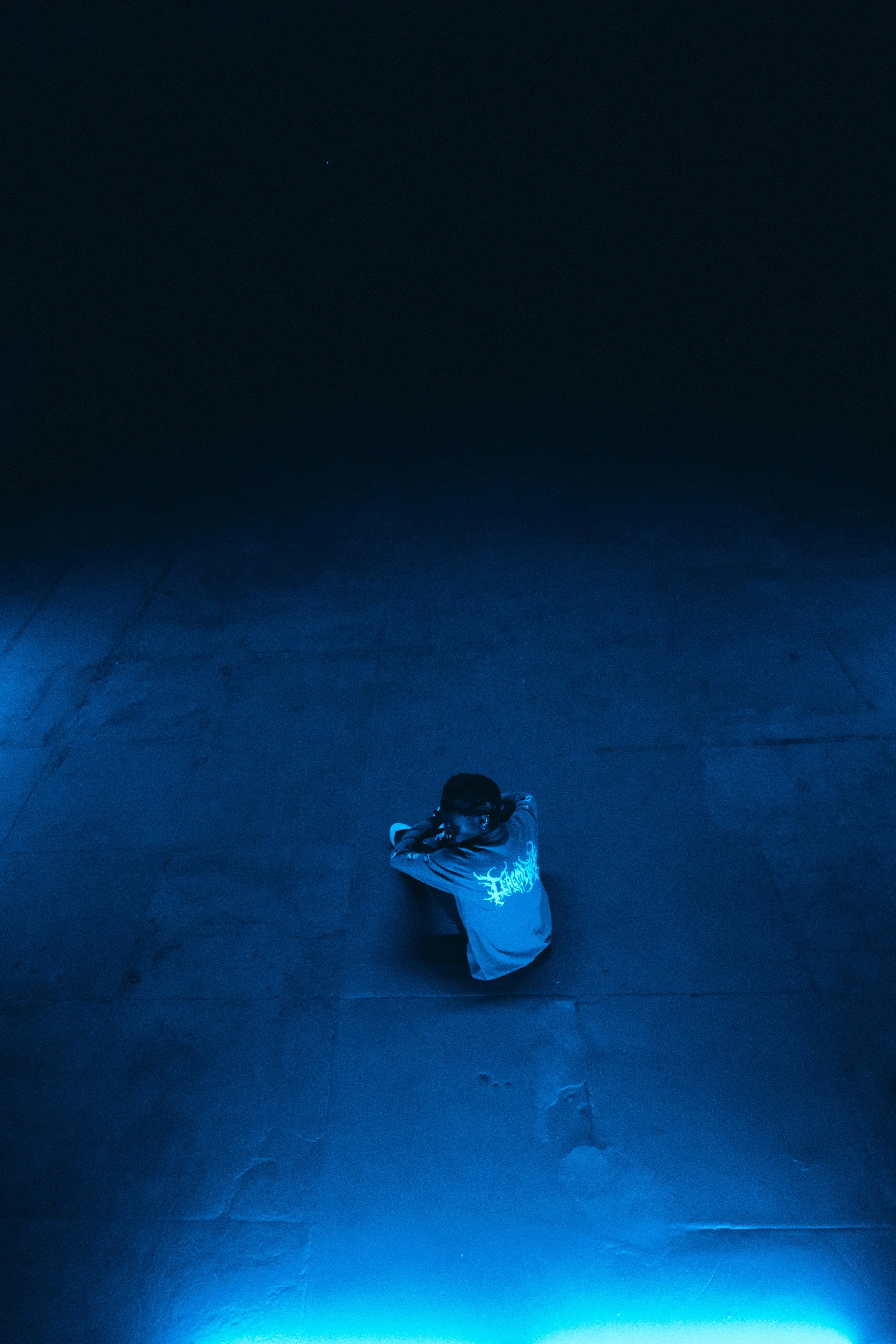 Person sitting on a dark floor illuminated by blue light, wearing a shirt with a graphic design on the back.