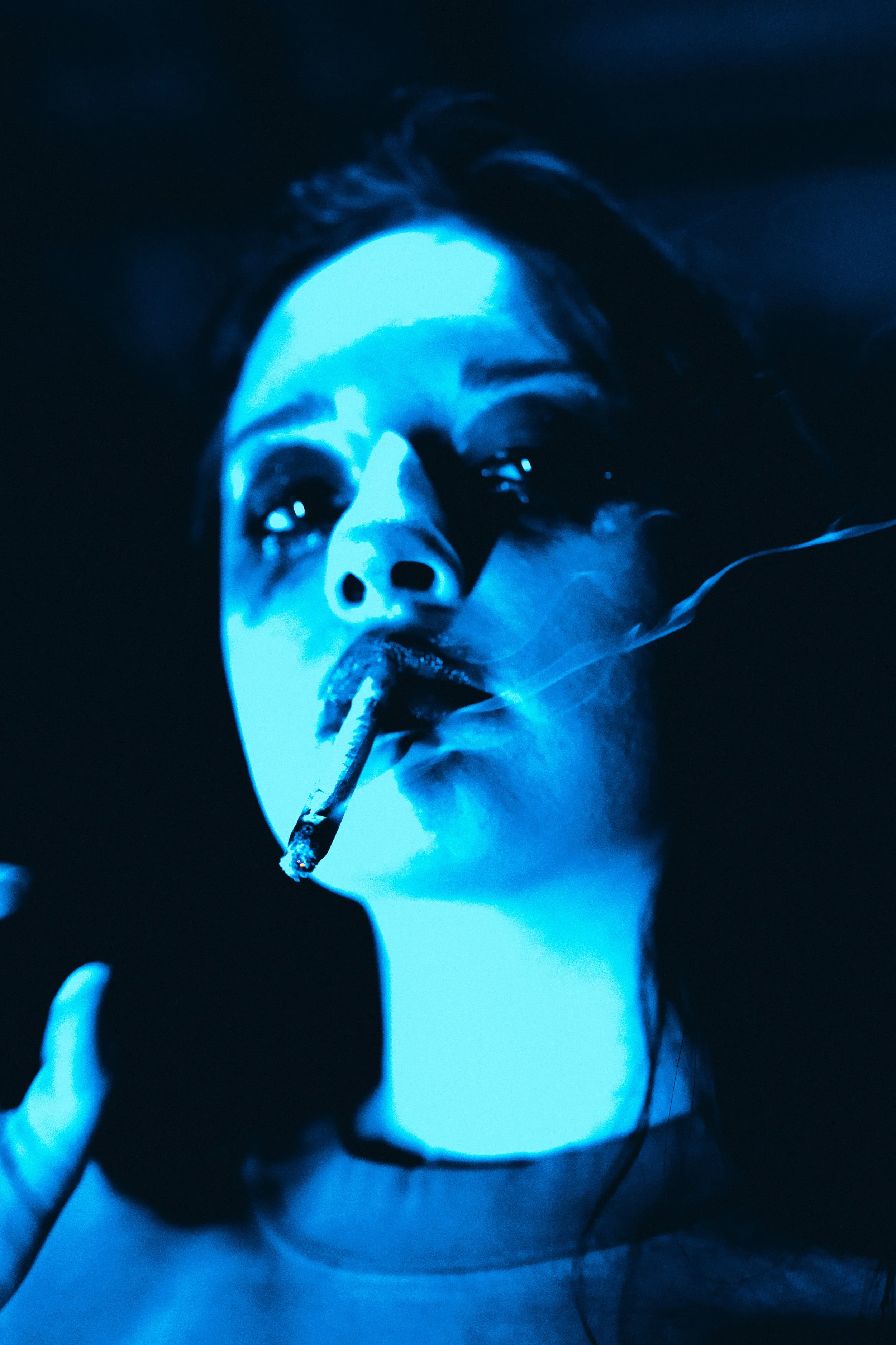 A person illuminated in blue light, holding a cigarette between their lips, with smoke rising.