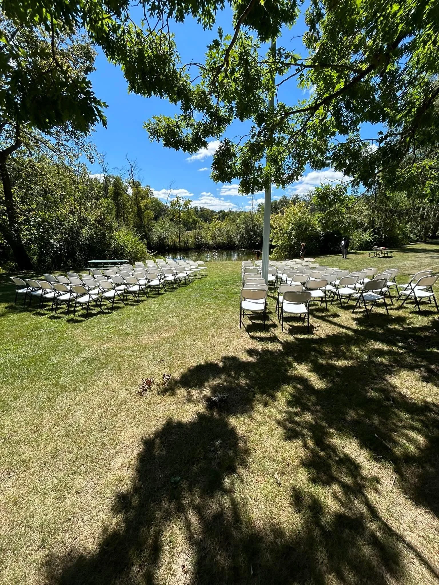 Outdoor wedding setup with white folding chairs on grass under trees, facing a wooded area with a blue sky and clouds.
