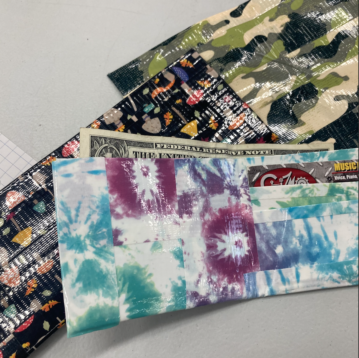 Duct tape wallets with various patterns including tie-dye, camouflage, and colorful designs, holding dollar bills and cards.