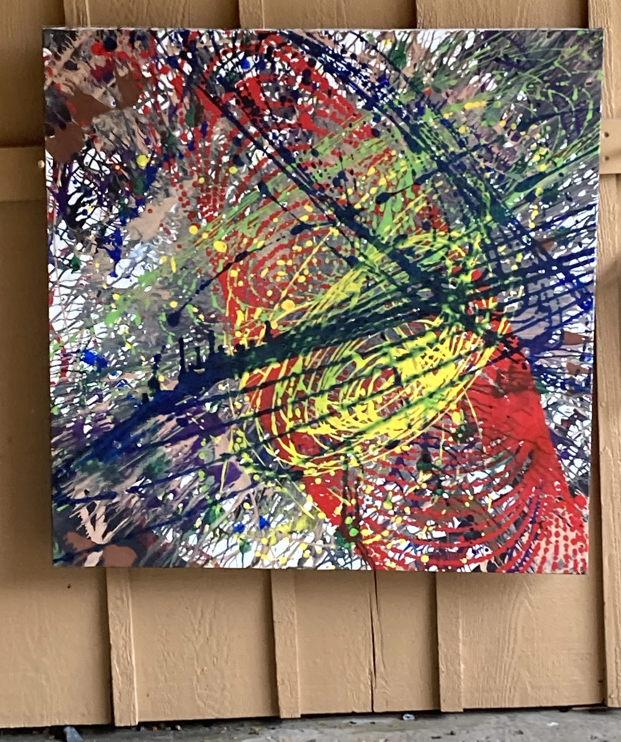 Abstract painting with splashes of red, green, yellow, blue, and black on a textured background leaning against a wooden wall.