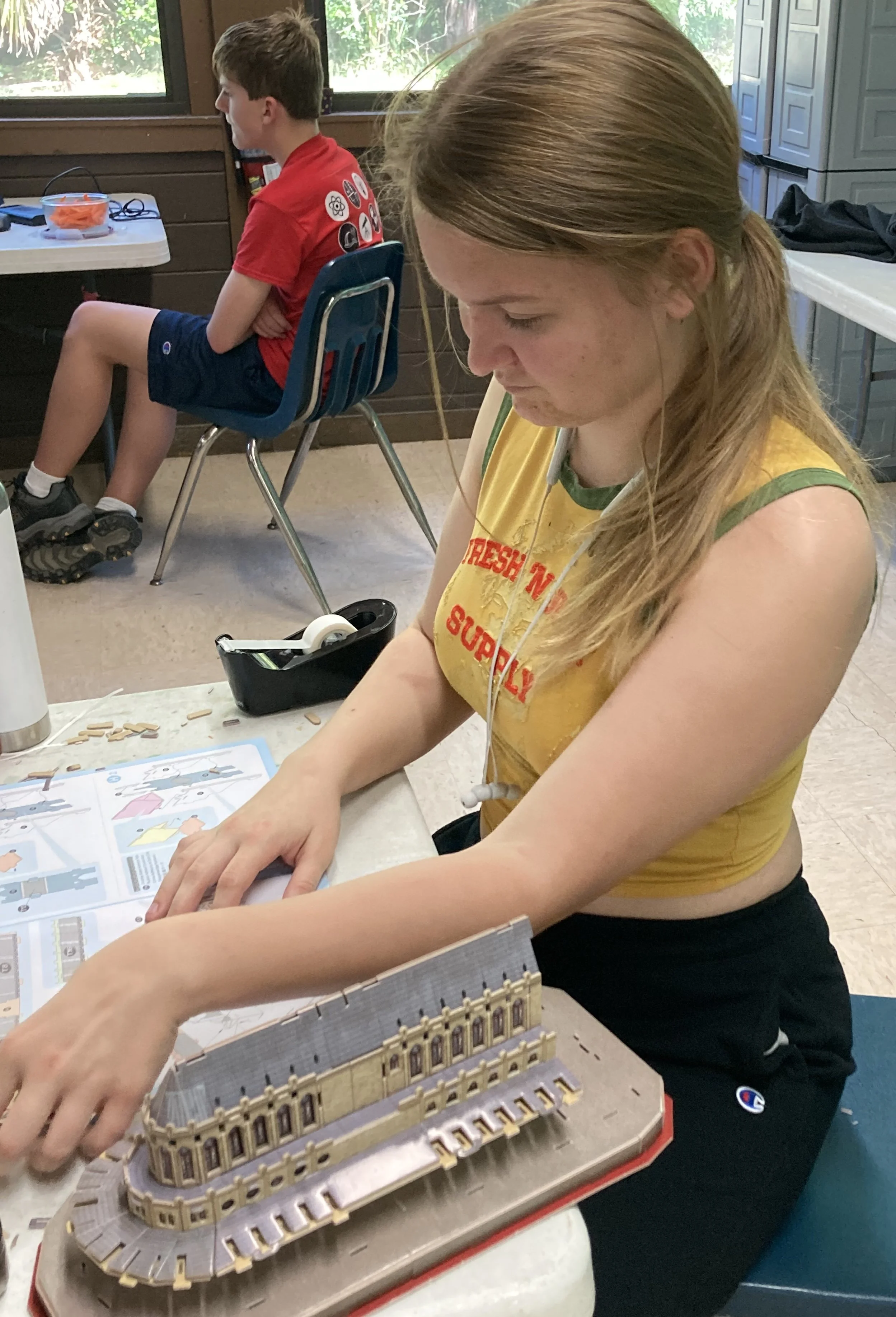 Young woman assembling a model building at a table, with a boy sitting in the background.