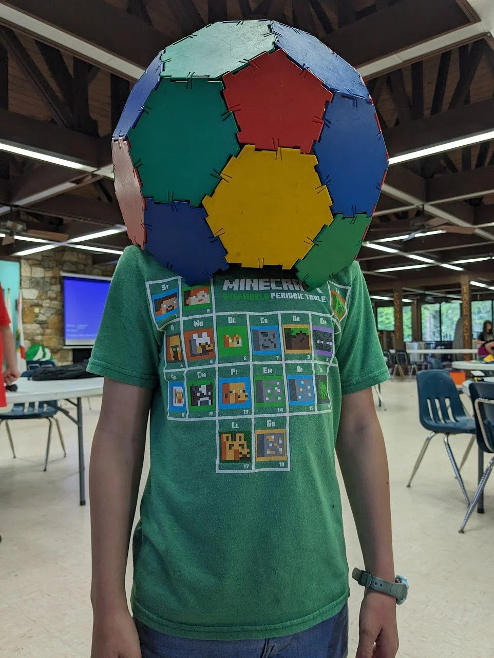Person wearing a large multicolored geometric shape on their head, standing indoors, wearing a green graphic T-shirt with pixelated designs, in a room with chairs and tables.