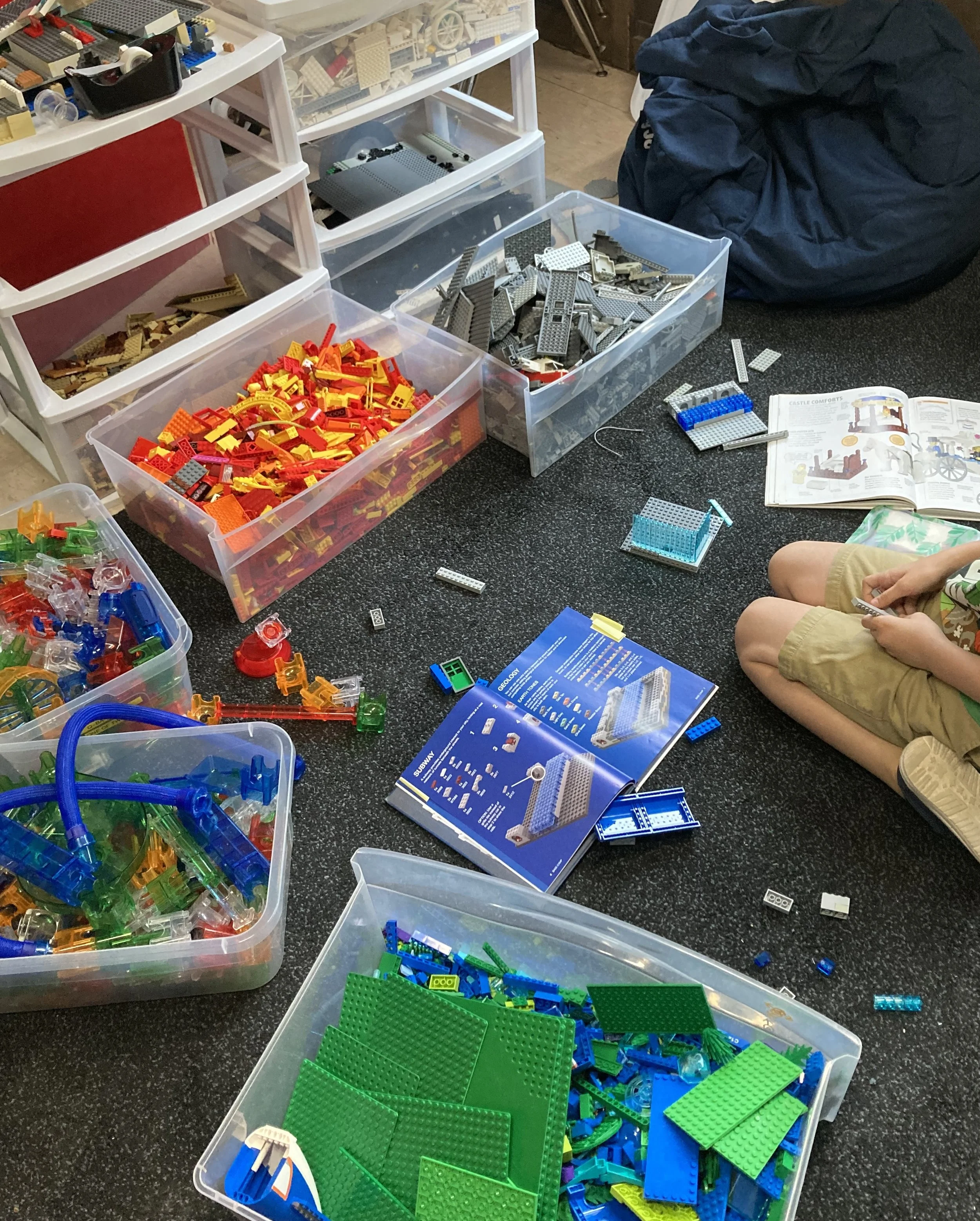 Various plastic containers filled with colorful Lego pieces and a child sitting nearby assembling them. An open instruction manual is visible on the floor.