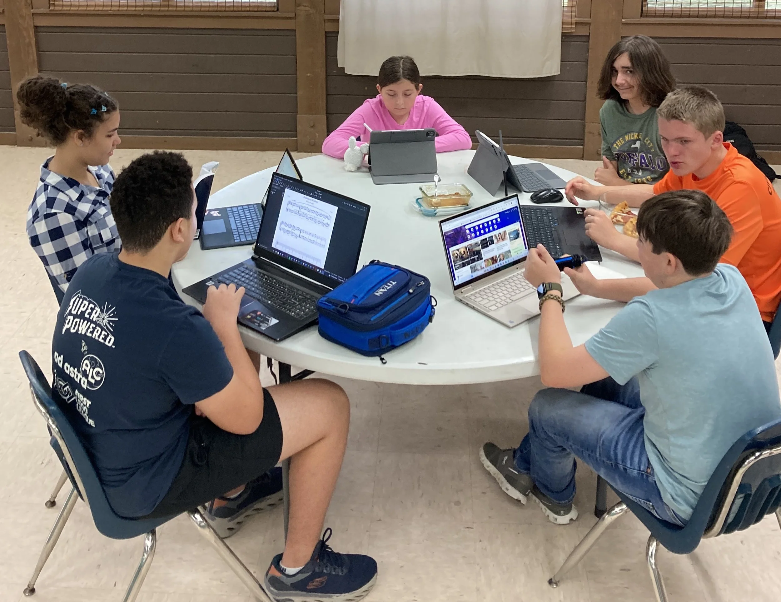 A group of six students sitting around a round table using laptops and tablets for a study session. They are focused on their screens, with one student writing and others viewing documents or online content. A snack and a blue lunch box are also on the table.