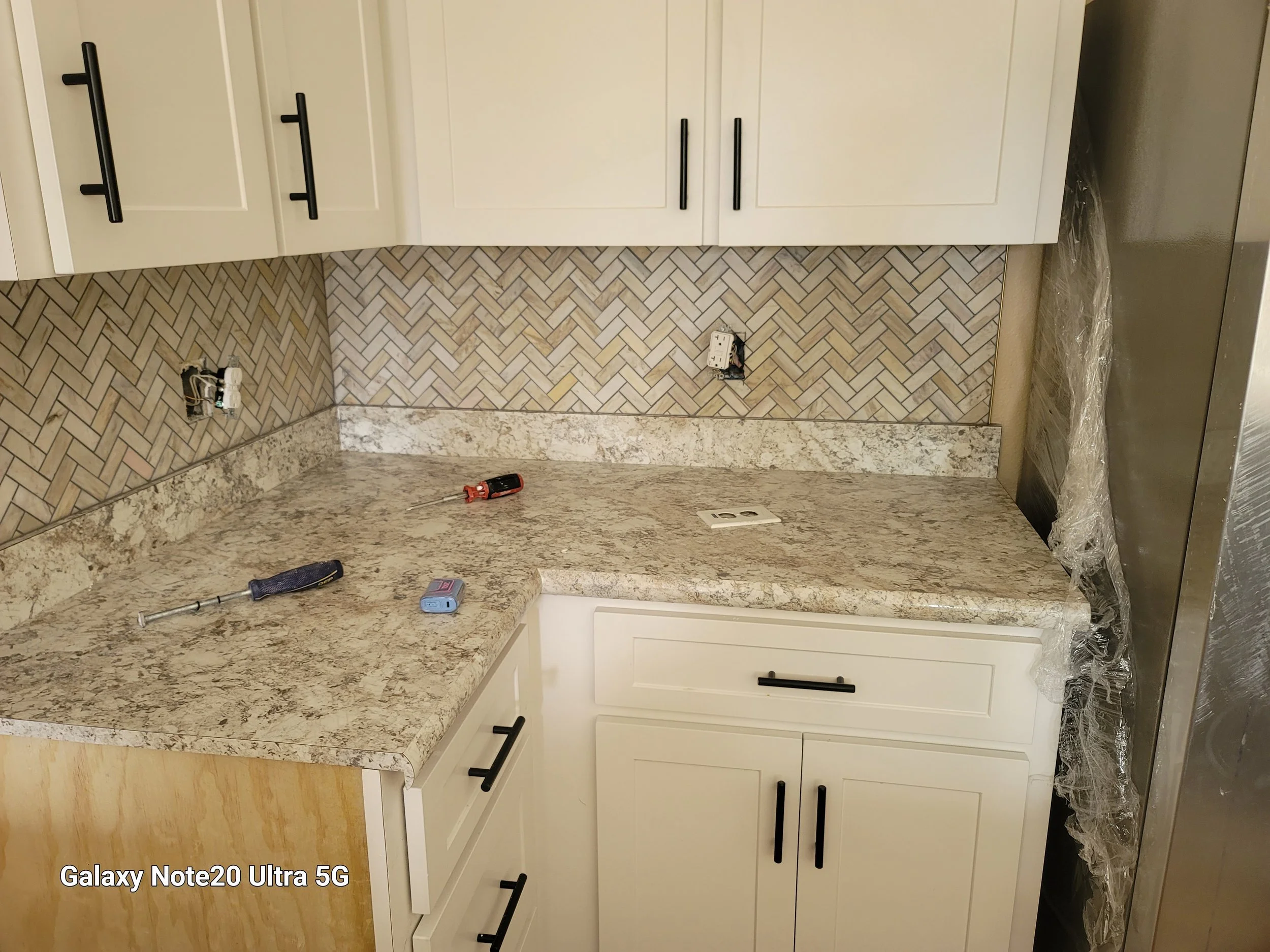 Kitchen countertop with tools and construction materials, including a screwdriver, a small level, and a screwdriver, with white cabinets and a patterned tile backsplash, showing work in progress.