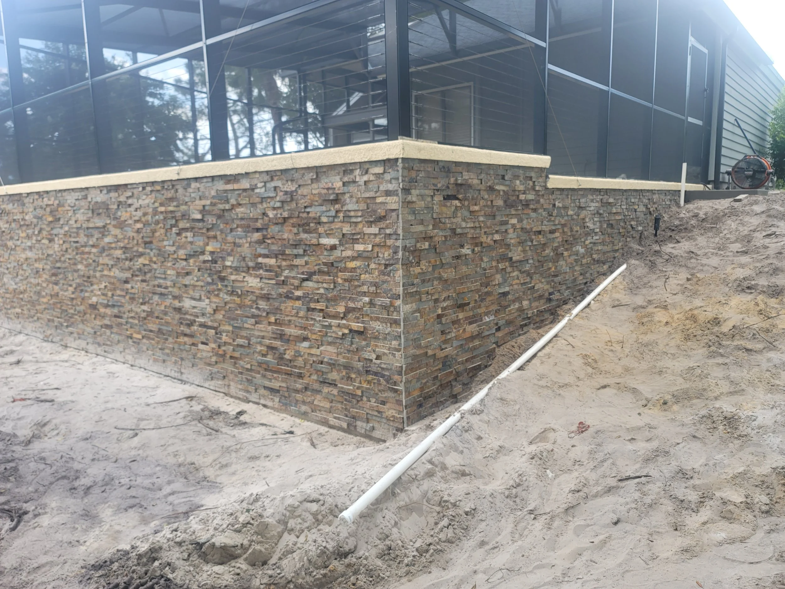 Close-up of a raised brick and stone retaining wall around a building with large glass windows, construction materials, and exposed soil in the foreground.
