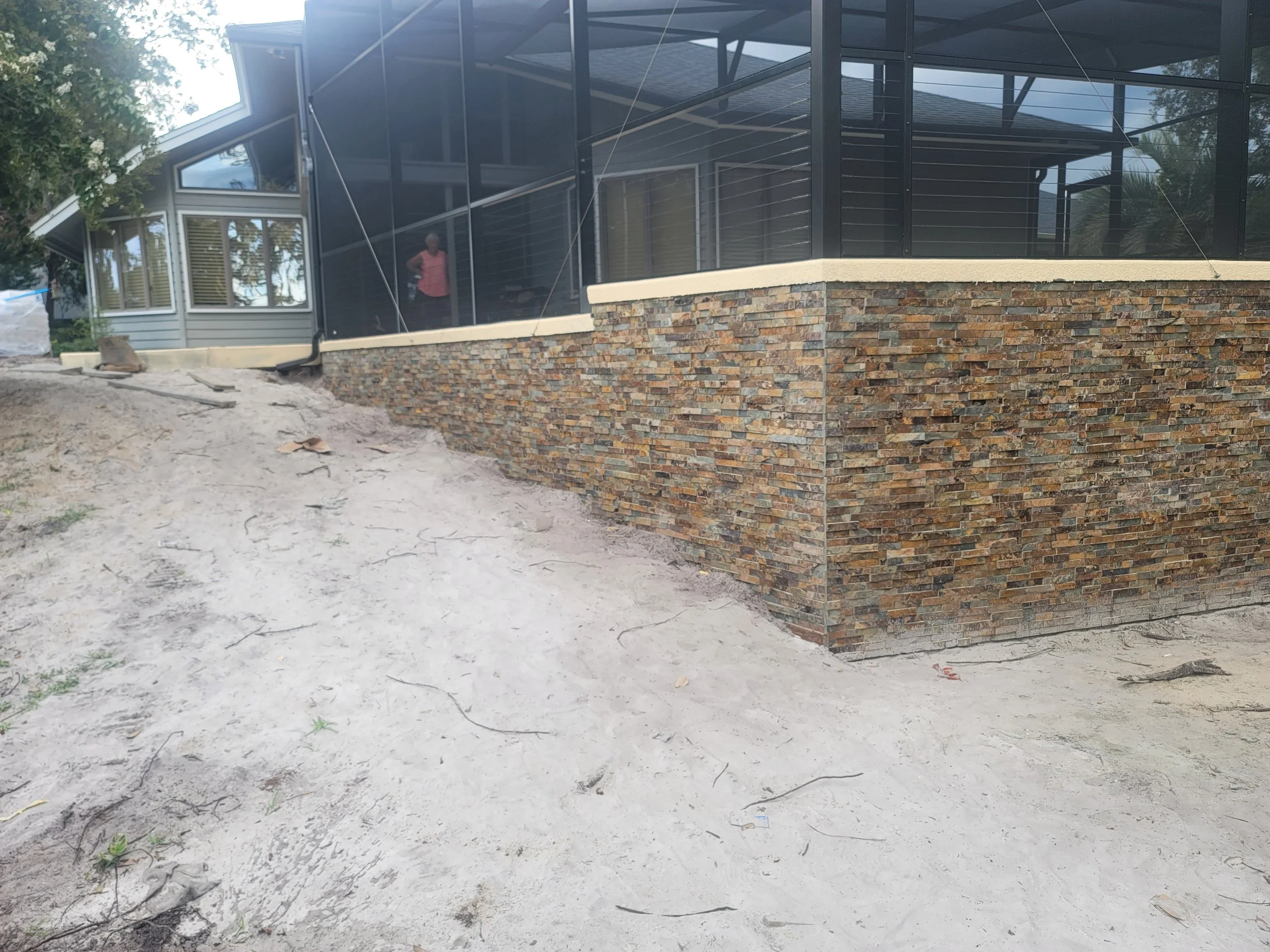 Exterior of a house under construction with a stone foundation, dirt ground with some small plants, and a screened porch with black framing.