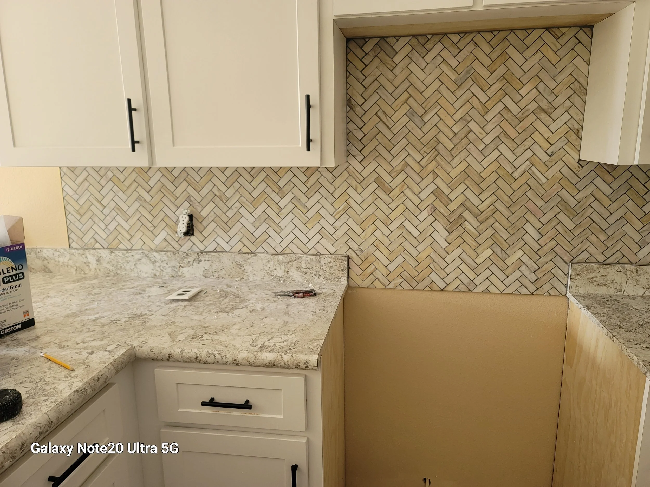 Kitchen corner with beige and cream-colored granite countertops, white cabinets with black handles, and a herringbone-patterned tile backsplash in shades of beige and brown.
