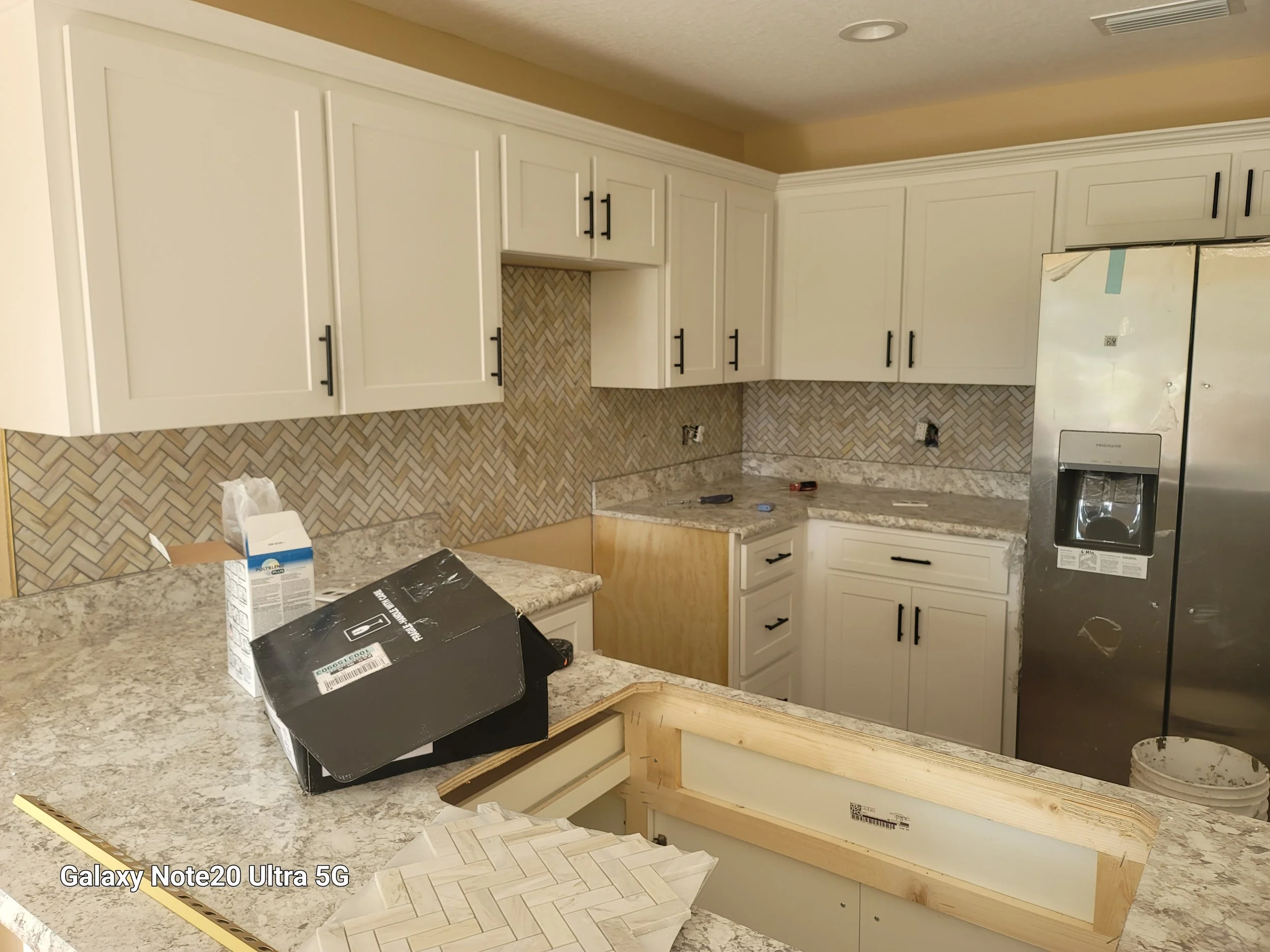 Kitchen with white cabinets, granite countertops, a stainless steel refrigerator, and a herringbone tile backsplash. Some tools and a box are on the counter, indicating ongoing renovation.