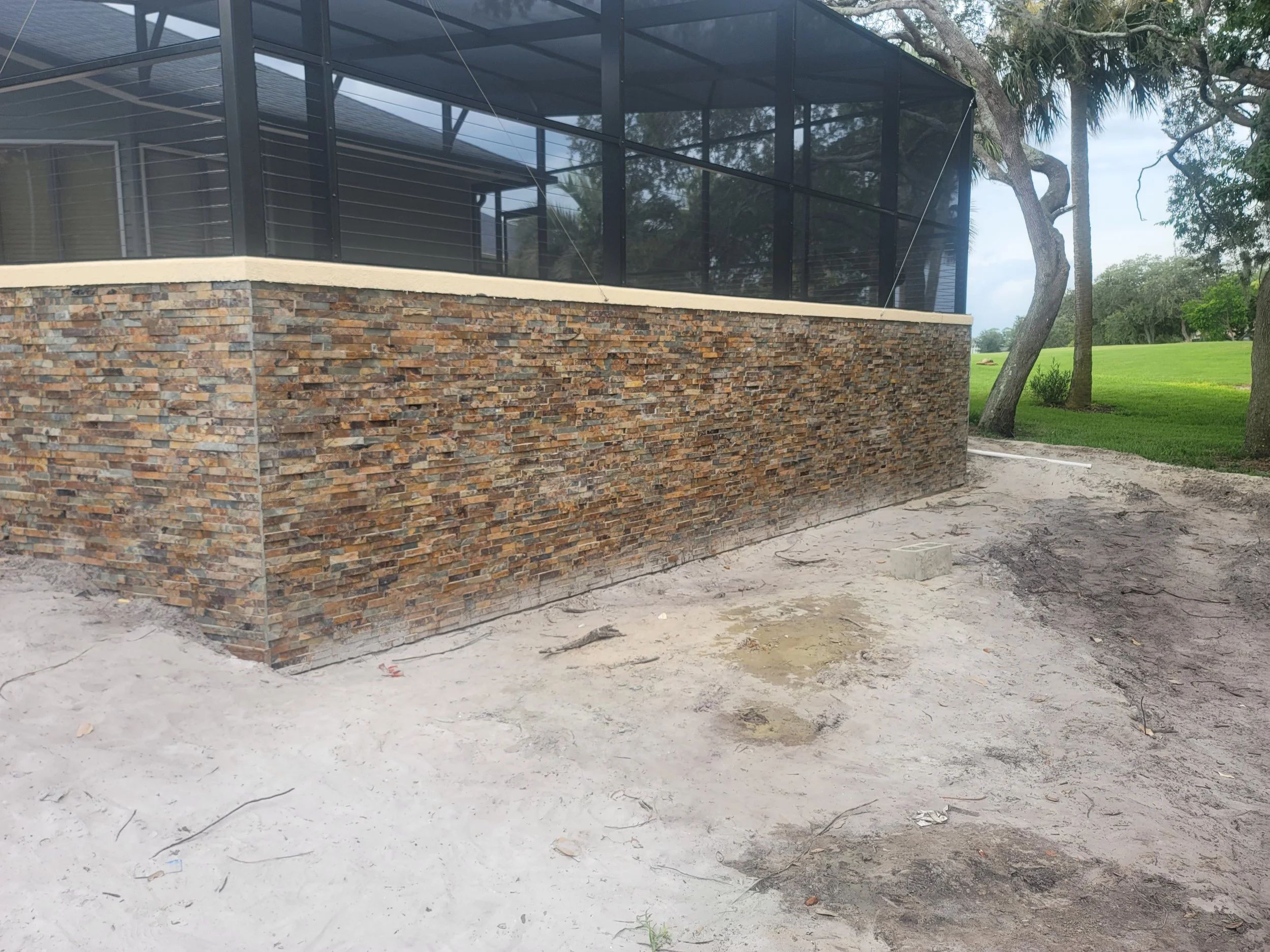 The image shows a corner of a house with a brick exterior and a screened porch, surrounded by sandy ground and green trees in the background.